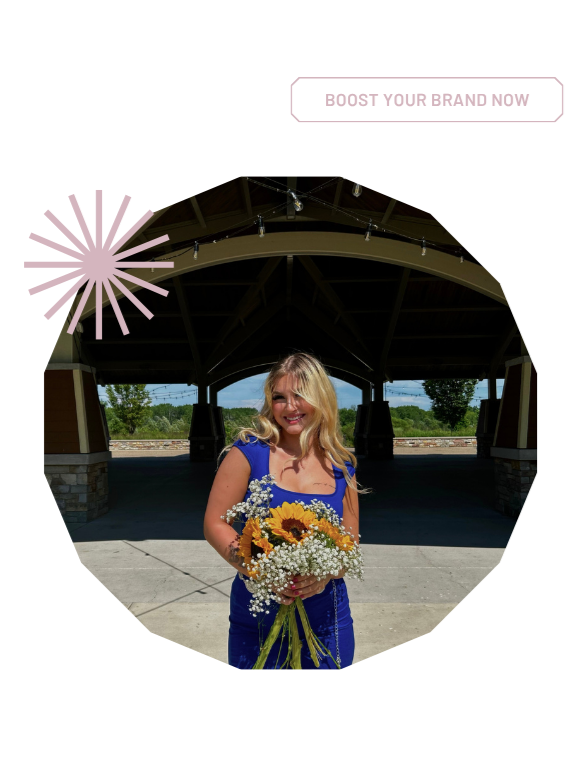A woman with blonde hair wearing a blue dress holding a bouquet of sunflowers and baby's breath, standing outdoors under a wooden pavilion with a sunny sky in the background.