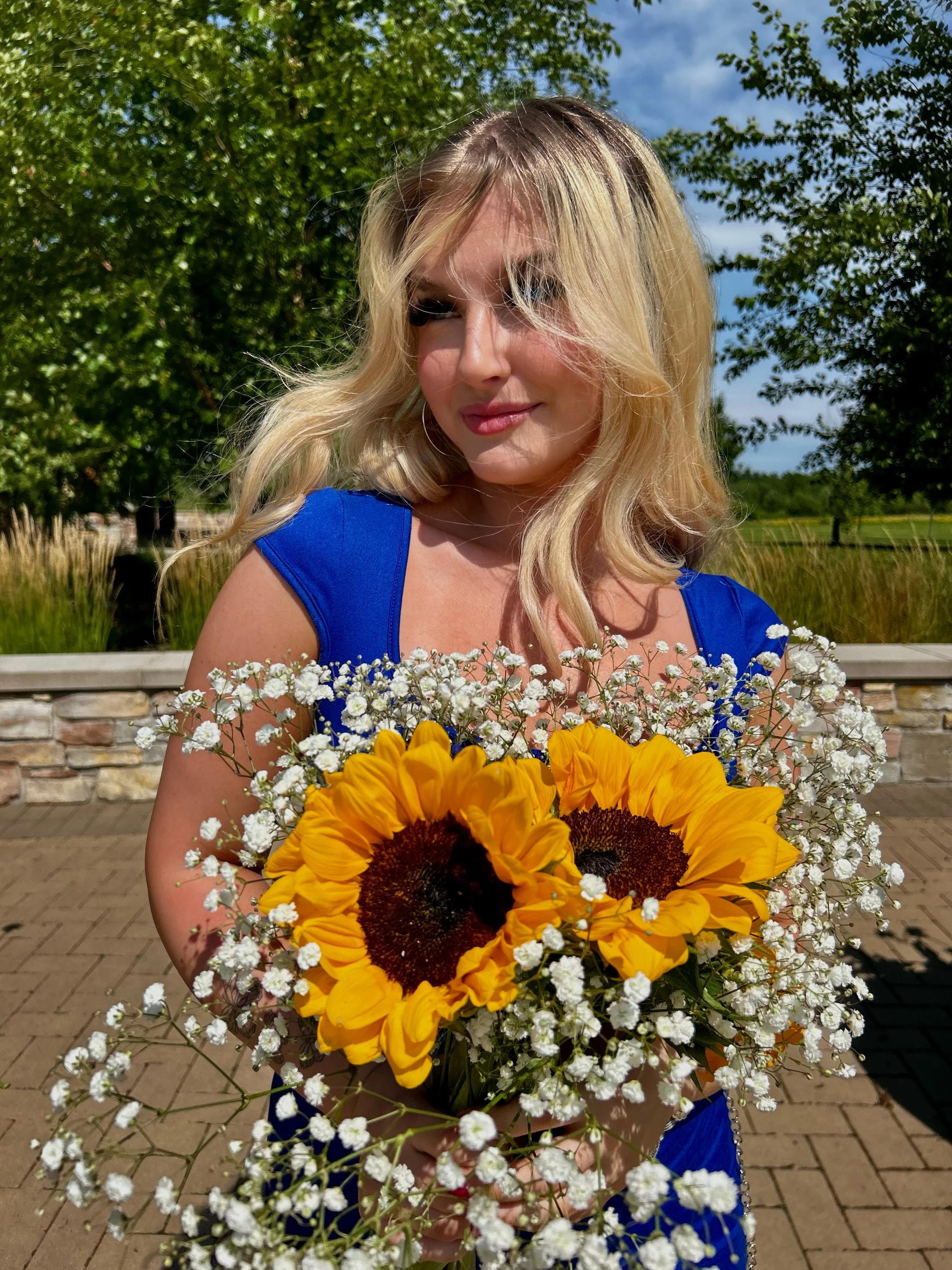 A woman with blonde hair wearing a blue dress holding a bouquet of sunflowers and small white flowers outdoors, with green trees and blue sky in the background.