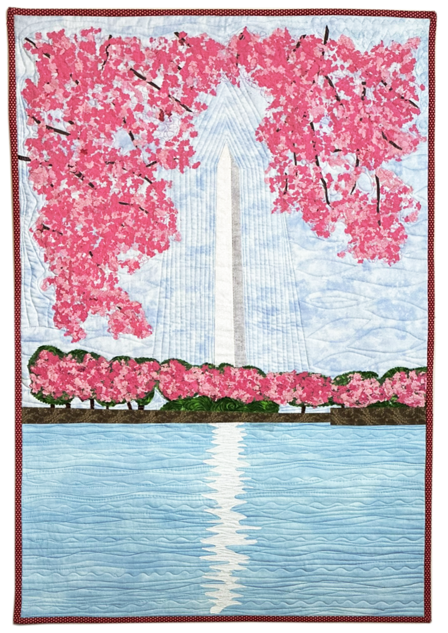 A quilted wall hanging depicting a landscape scene with pink cherry blossom trees, a river, and a blue sky.