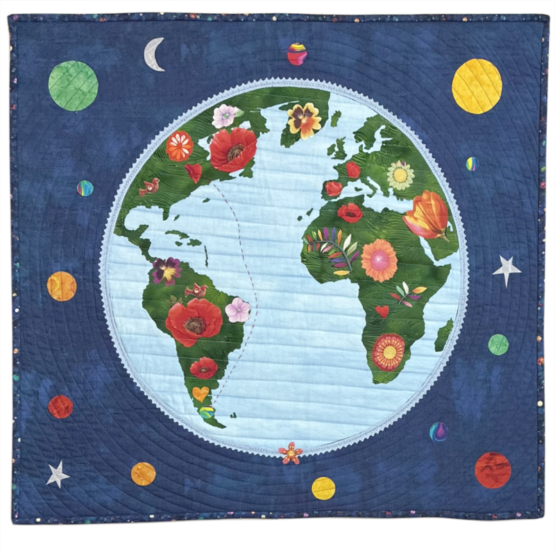 Fabric quilt featuring a stylized world map with continents decorated with colorful flowers, surrounded by a dark blue background with planets, stars, and moon motifs.