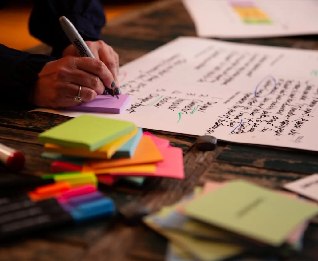 A person writing on a purple sticky note with a marker on a wooden table. Several sheets of paper with handwritten notes, a stack of brightly colored sticky notes, and various highlighters are also on the table.