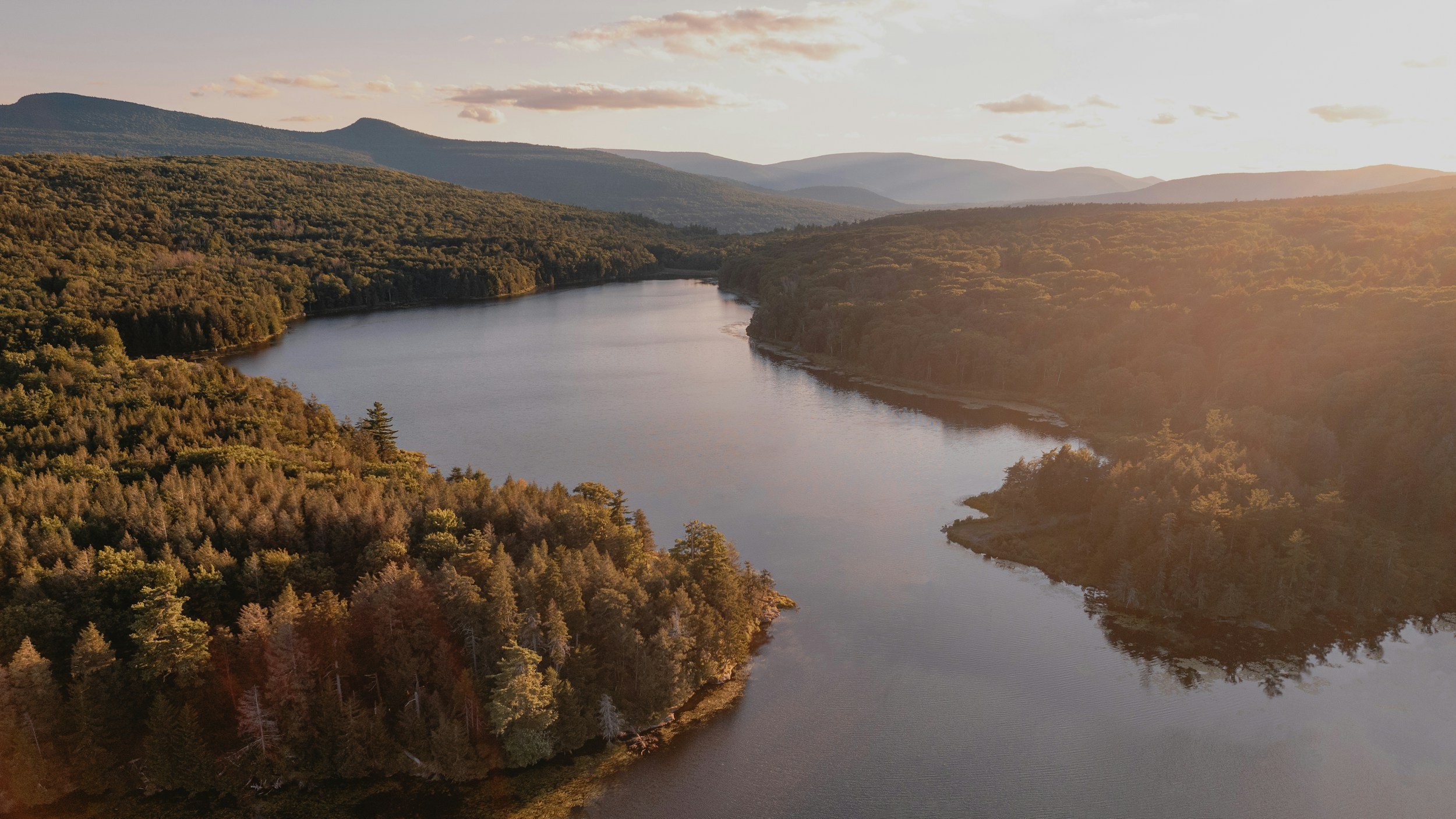 A landscape view of a river winding through a forested valley with mountains in the background during sunset.