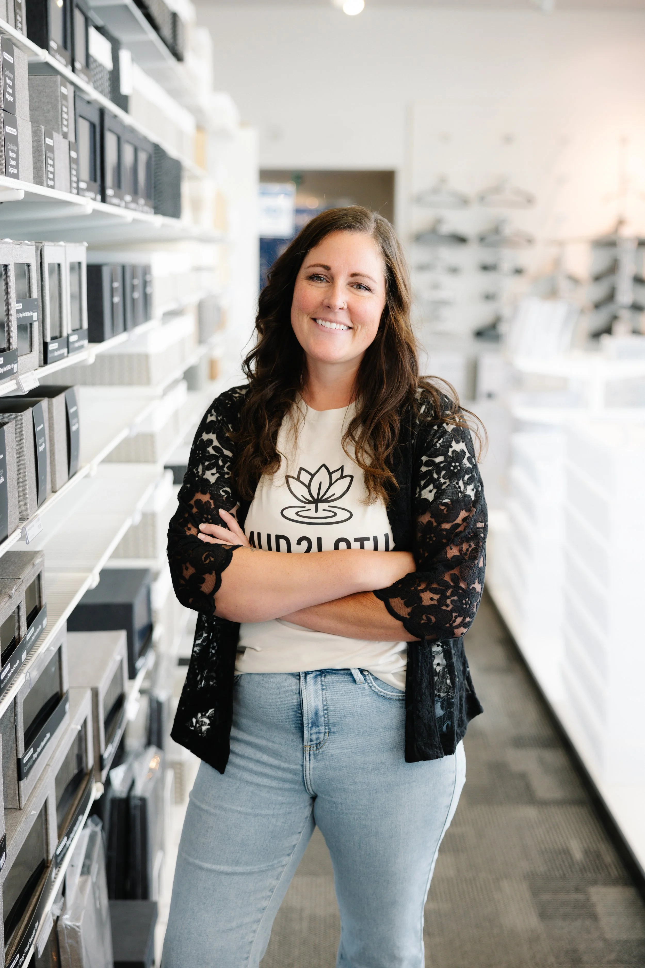 A woman with long brown hair smiling and crossing her arms, standing in a store aisle with shelves of small storage boxes around her, wearing a white t-shirt with a lotus flower design and the word 'MULTICULTURAL,' and a black lace cardigan.
