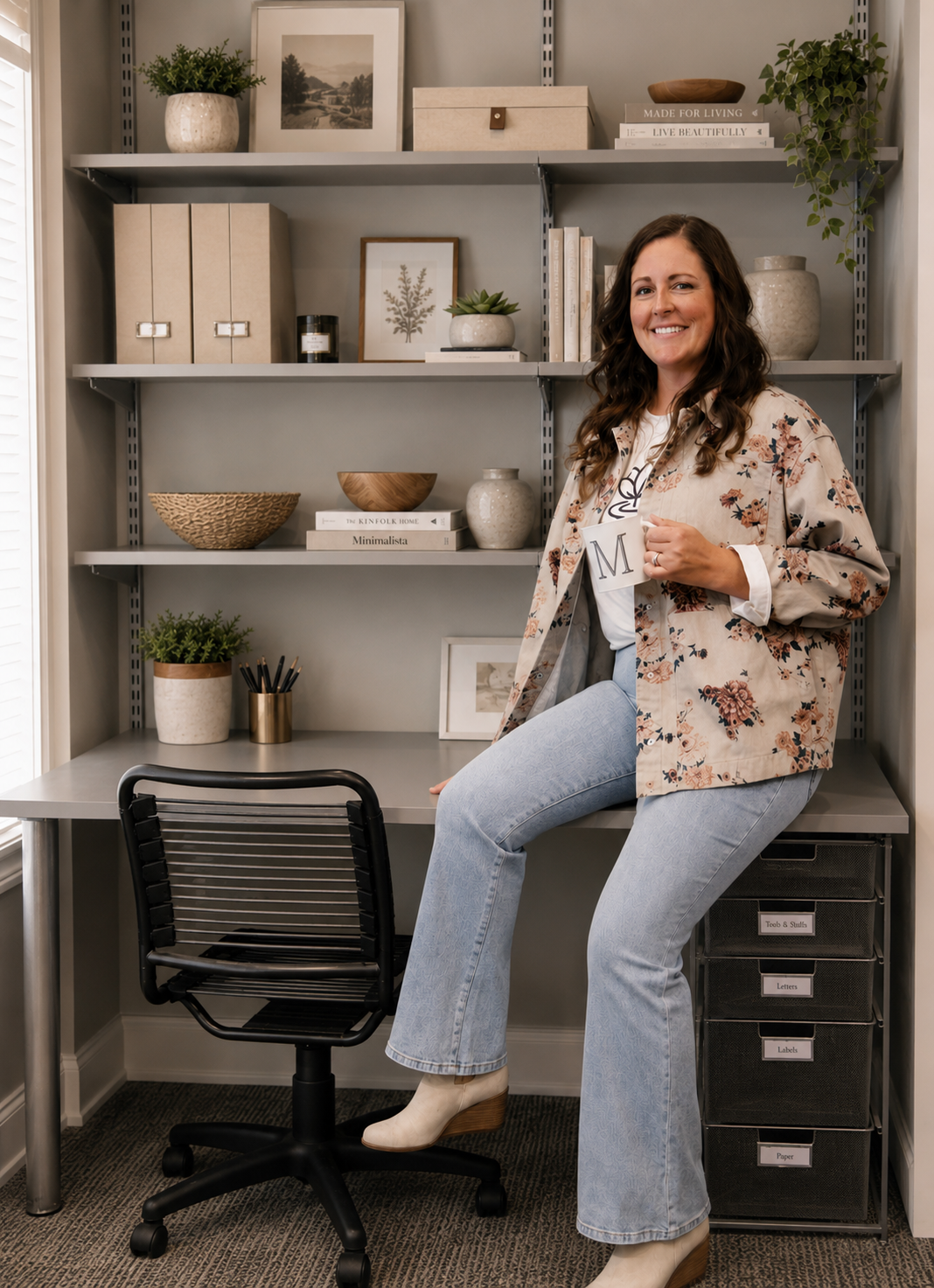 A woman sitting on a desk in a home office, holding a mug with the letter "M", smiling, with a bookshelf behind her filled with decorative items, books, and framed artwork.