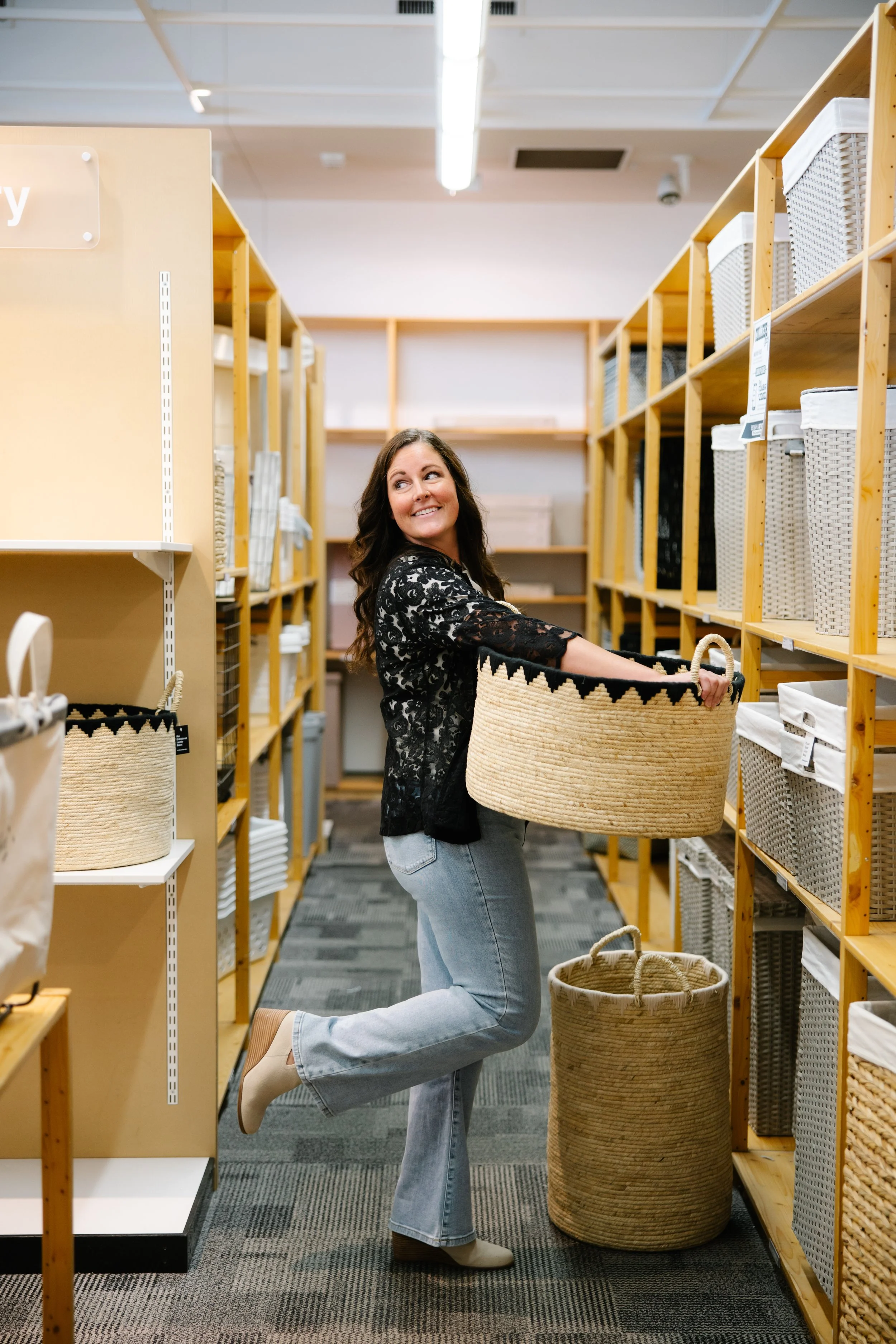 A woman with long brown hair kneeling on one knee in a store aisle, holding a large woven basket with a black-patterned rim, surrounded by shelves with similar baskets and storage containers.