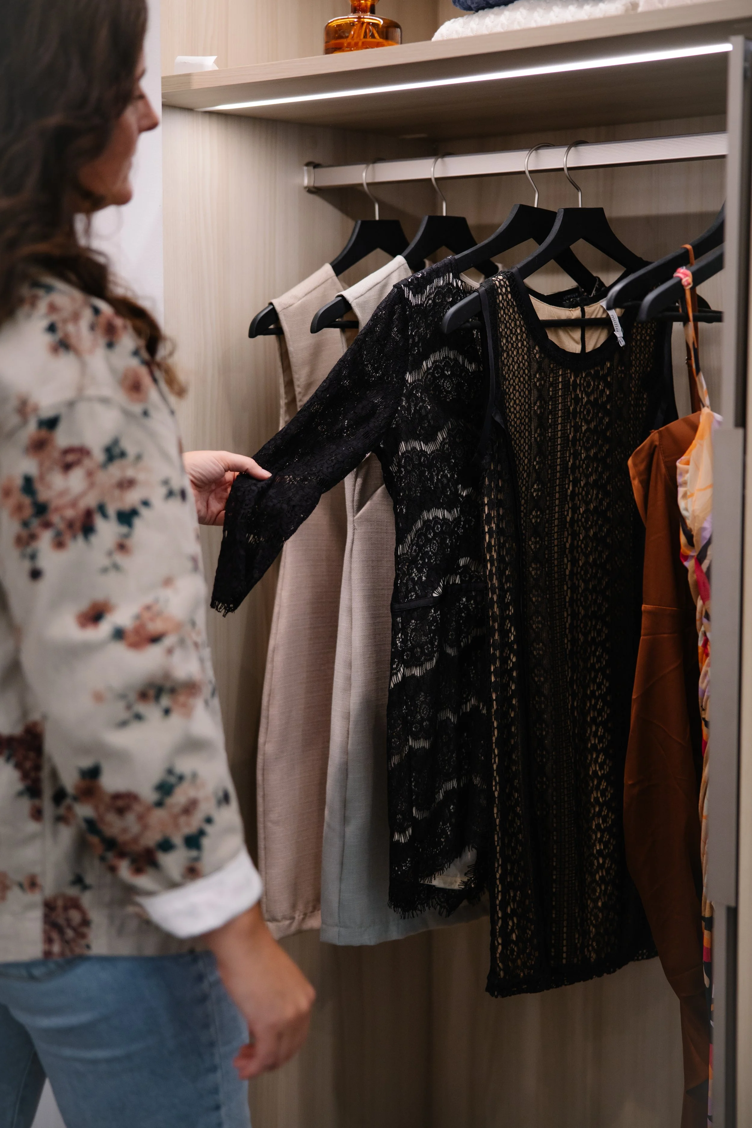 A woman with long, wavy hair wearing a floral blouse looks at black lace and beige dresses hanging on a rod in a clothing store fitting room.