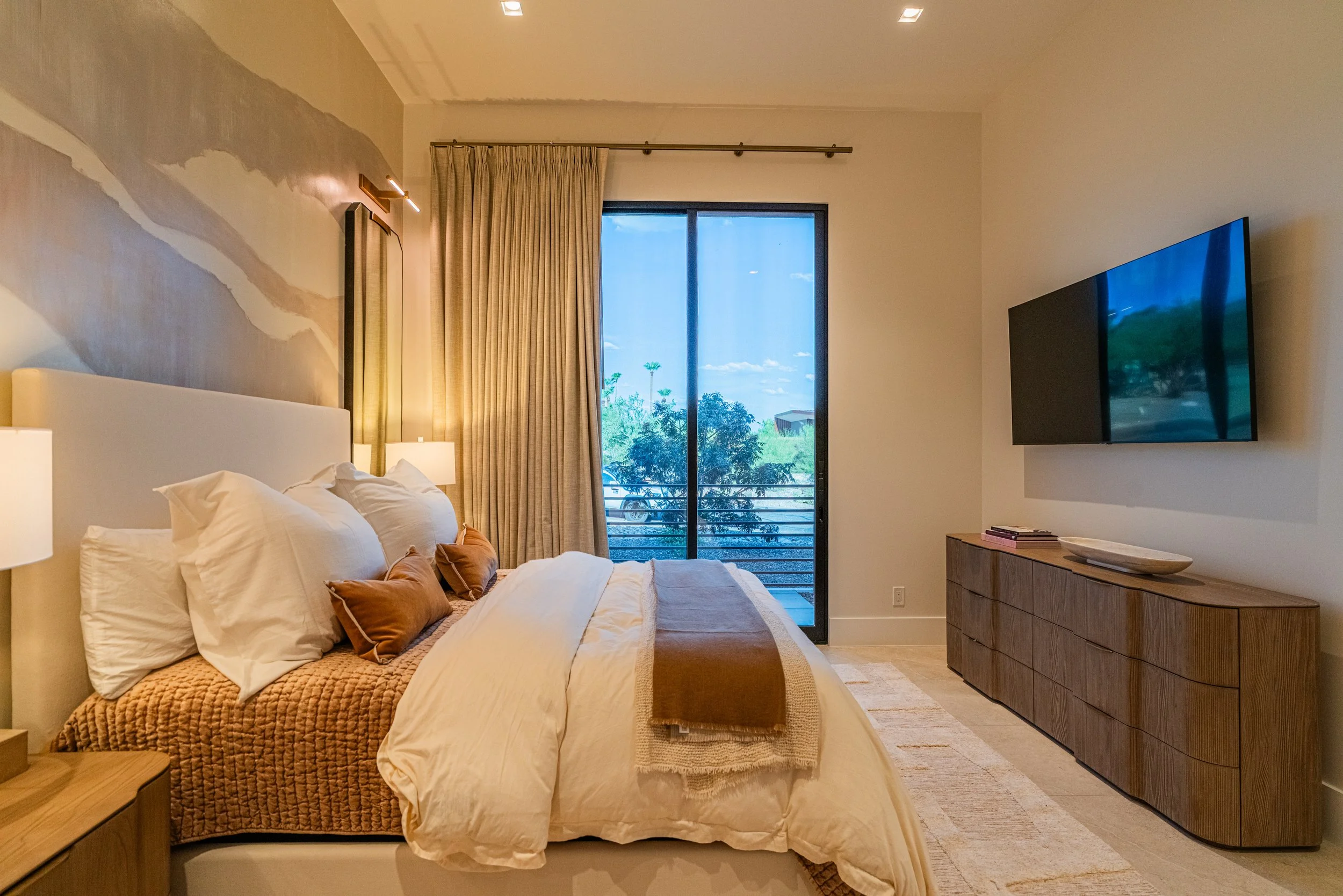 A modern bedroom with a large bed, white bedding, and decorative pillows, a wooden side table, a dresser, a wall-mounted flat screen TV, a sliding glass door with beige curtains, and outdoor greenery visible through the window.