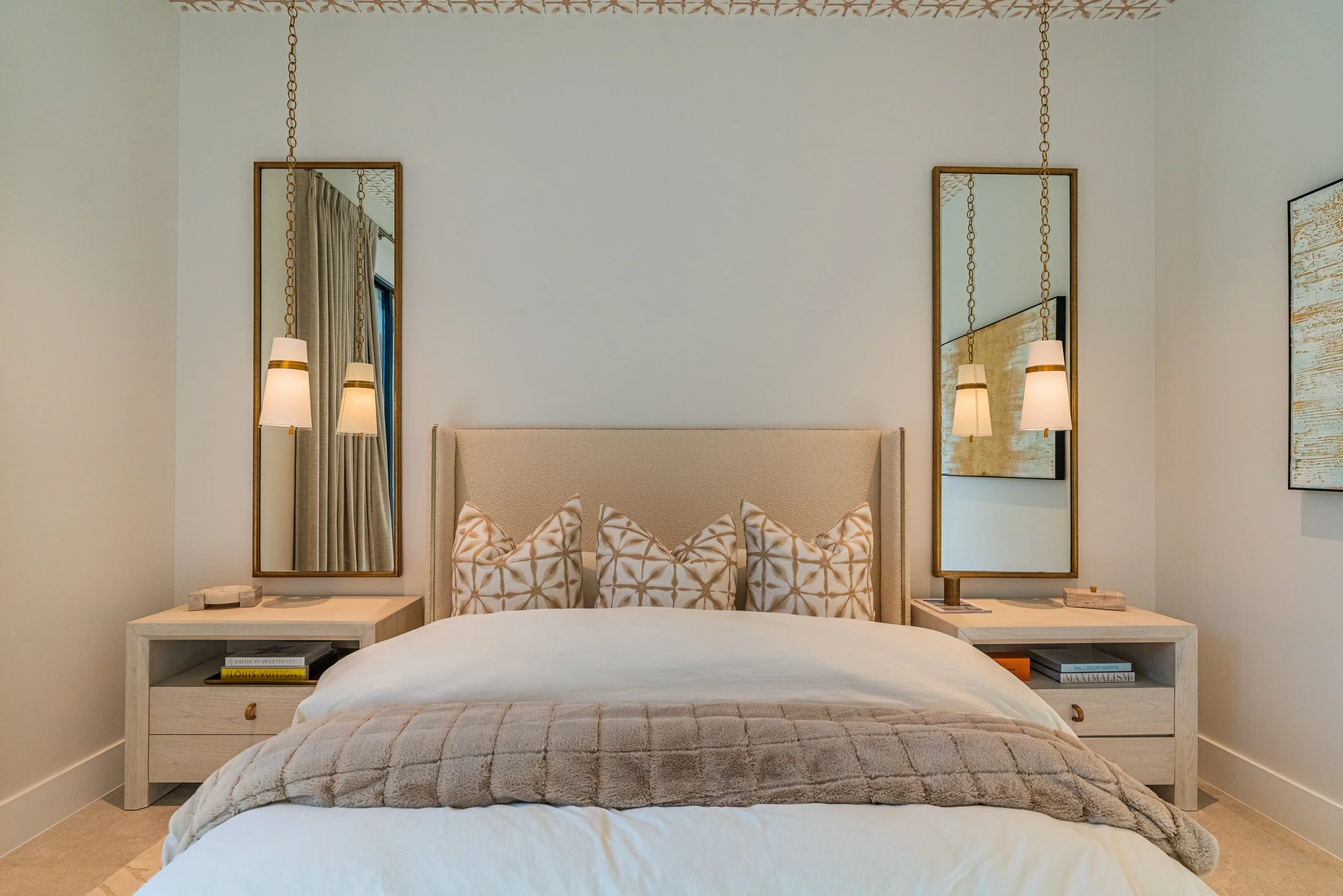 Modern bedroom with a beige upholstered headboard, decorative pillows, and matching bedside tables with lamps and books. Mirrors and artwork are mounted on the wall above the bed.
