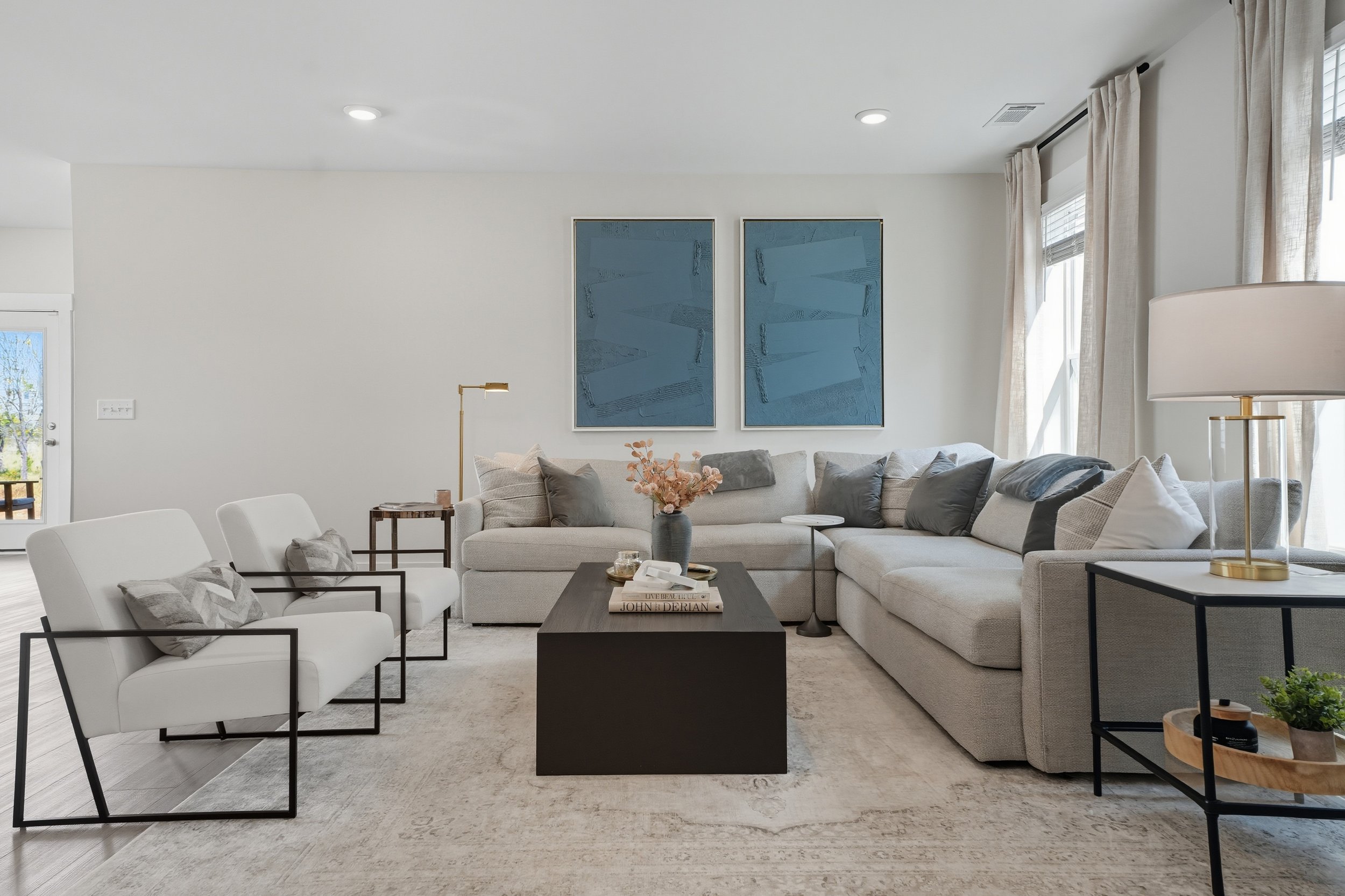 Modern living room with light-colored sofa, white armchairs, black coffee table, decorative books, pink flowers, artwork on wall, large windows with curtains, and a floor lamp.