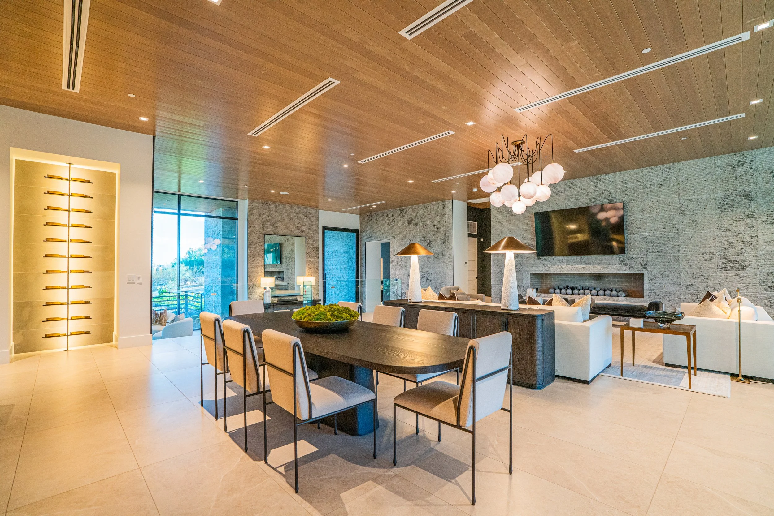 A modern living room with a wooden ceiling, a large wall-mounted TV, a beige sofa, a fireplace with decorative stones, and a black dining table with white chairs. There are large windows and artistic lighting fixtures.