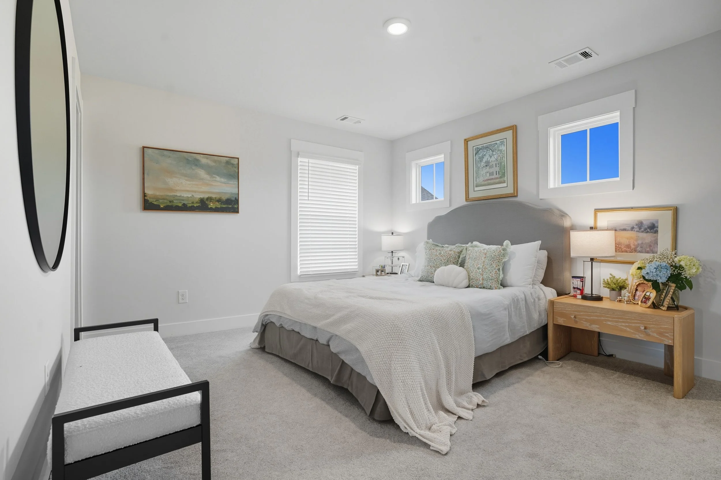 A bright bedroom with white walls, a bed with a white bedspread and decorative pillows, a wooden nightstand with a lamp, framed artwork, and windows showing blue sky.