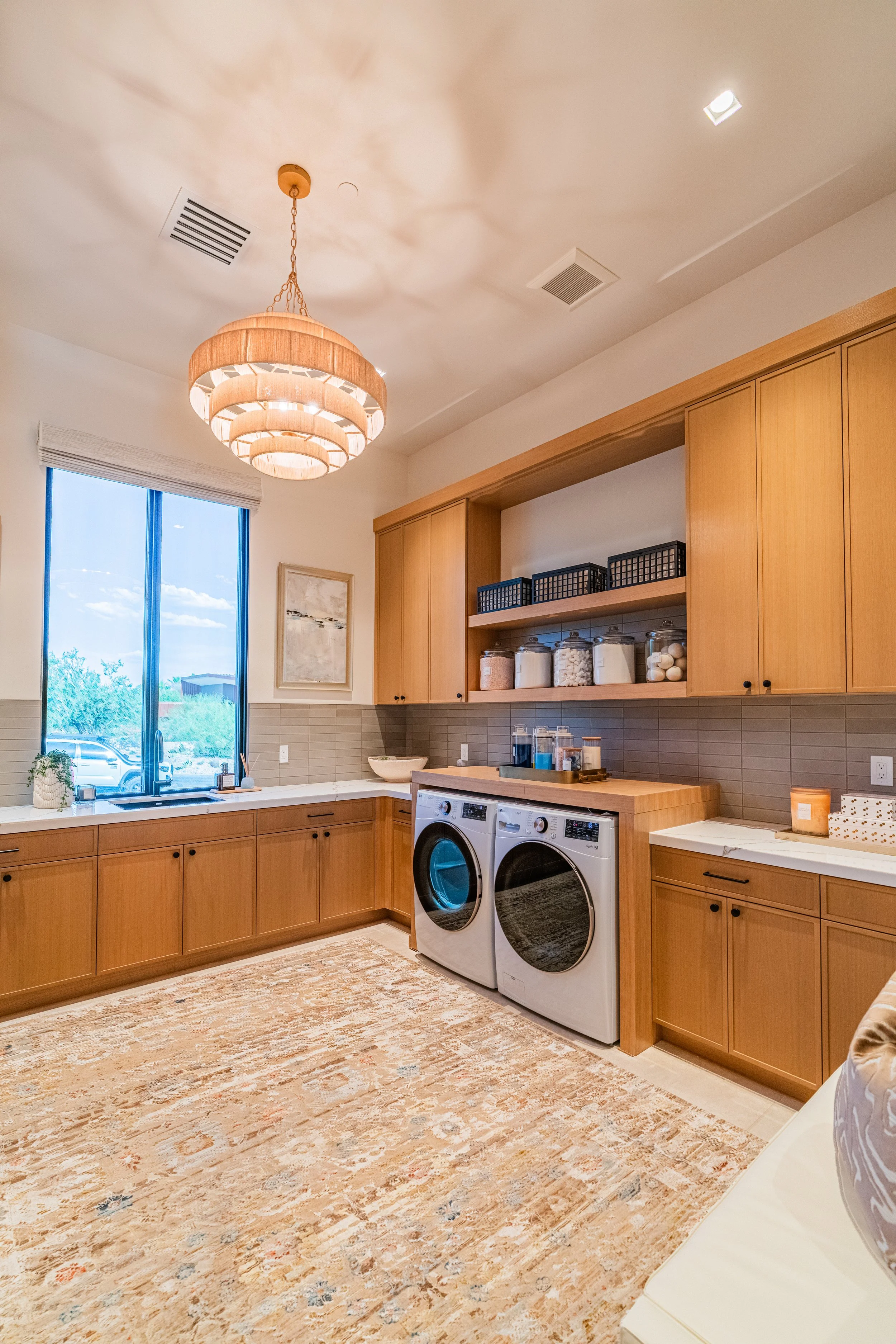 A cozy laundry room with natural wood cabinets, a washer and dryer, a window letting in natural light, a chandelier ceiling light, decorative jars on a shelf, and a textured beige rug on the floor.