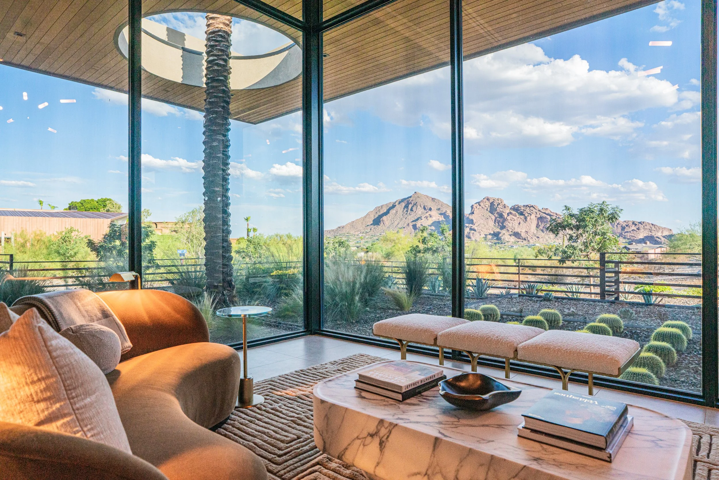 Living room with large floor-to-ceiling windows looking out at desert landscape with mountains, trees, and blue sky, contemporary furniture, marble coffee table, books, and potted plants.