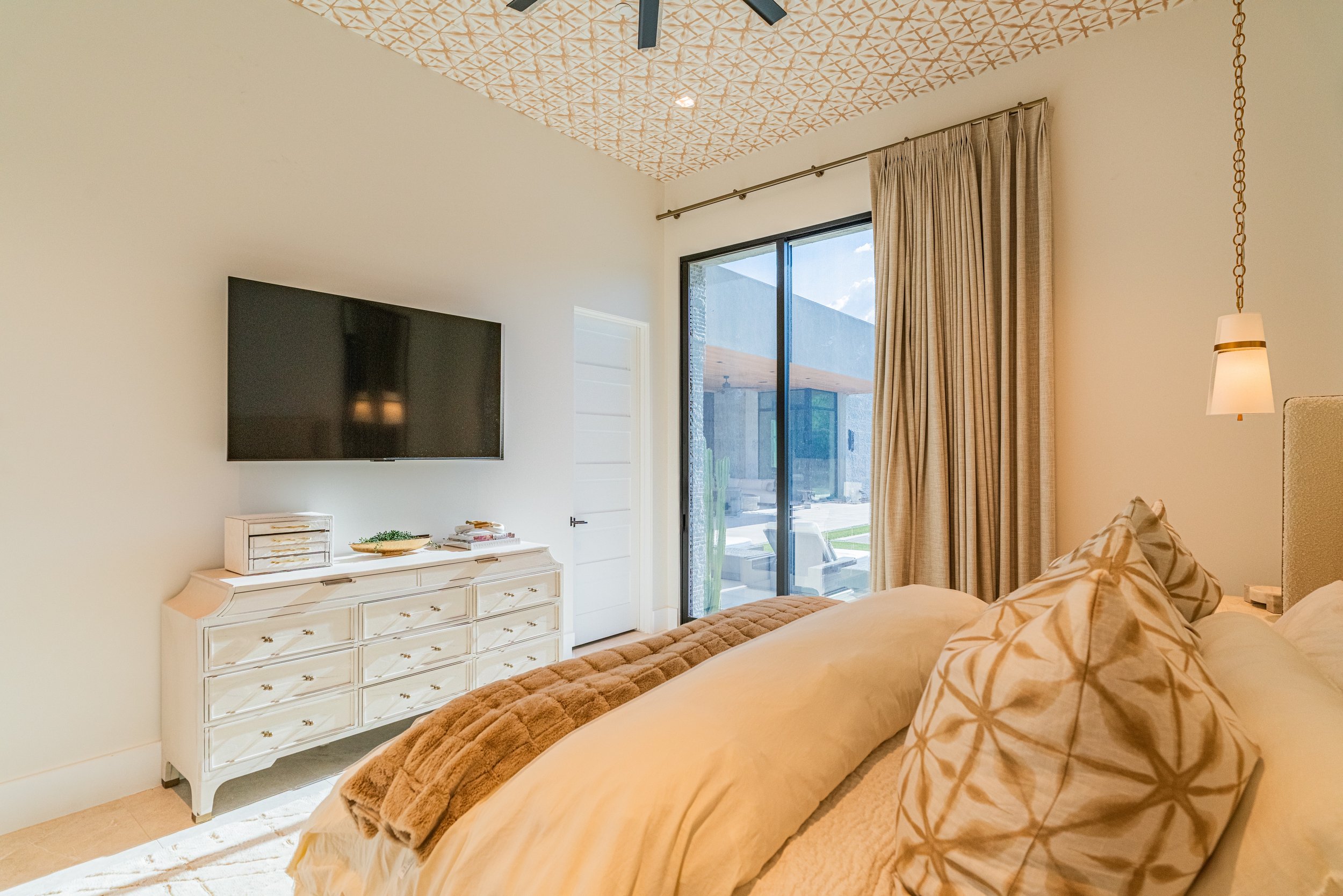 A bedroom with a large window covered by beige curtains, a wall-mounted TV, a white dresser with drawers, and a beige bed with patterned pillows. There is a pendant light hanging from the ceiling. The outdoors visible through the window shows a patio