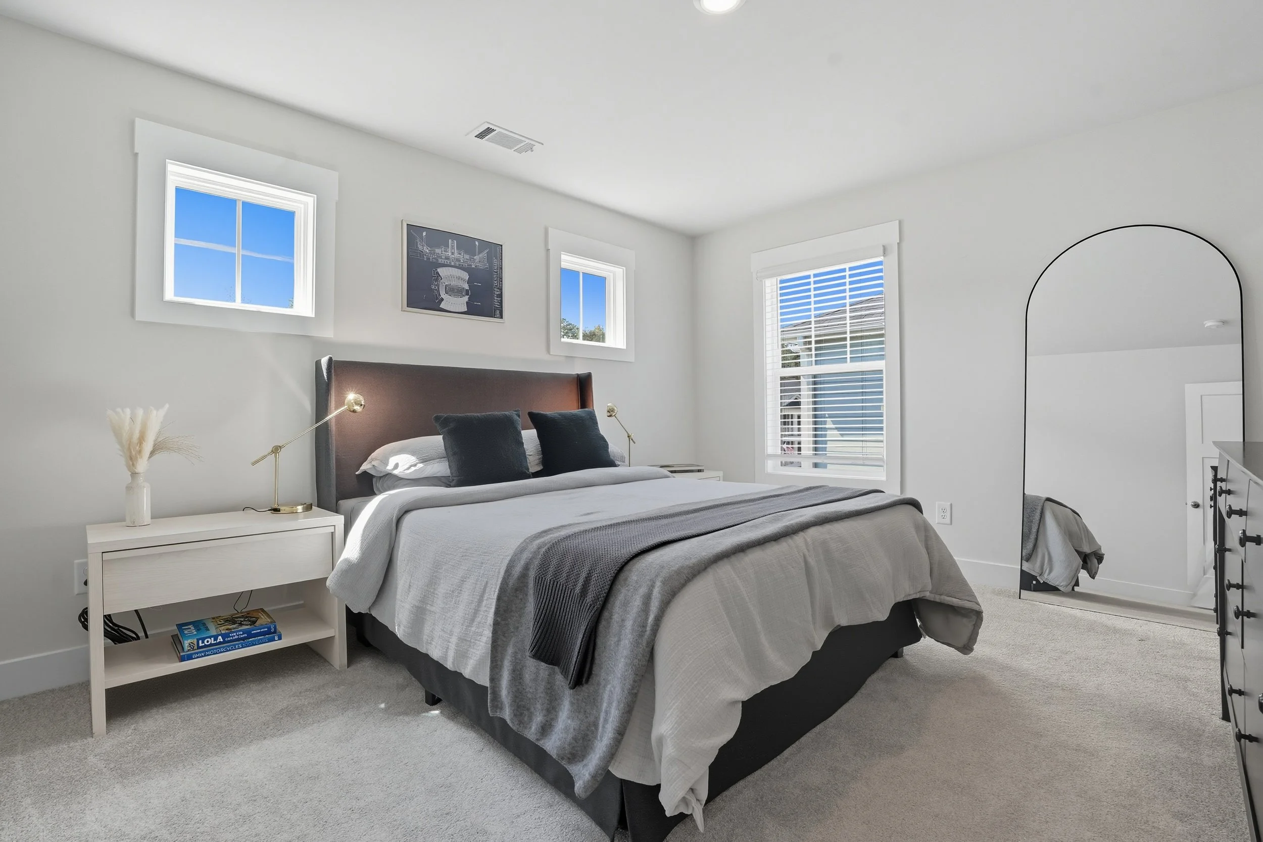 A modern bedroom with white walls and ceiling, two small windows with white trim, and a large bed with white and gray bedding and dark pillows. There is a white nightstand with a modern gold lamp, a vase with pampas grass, and a book. A black dresser