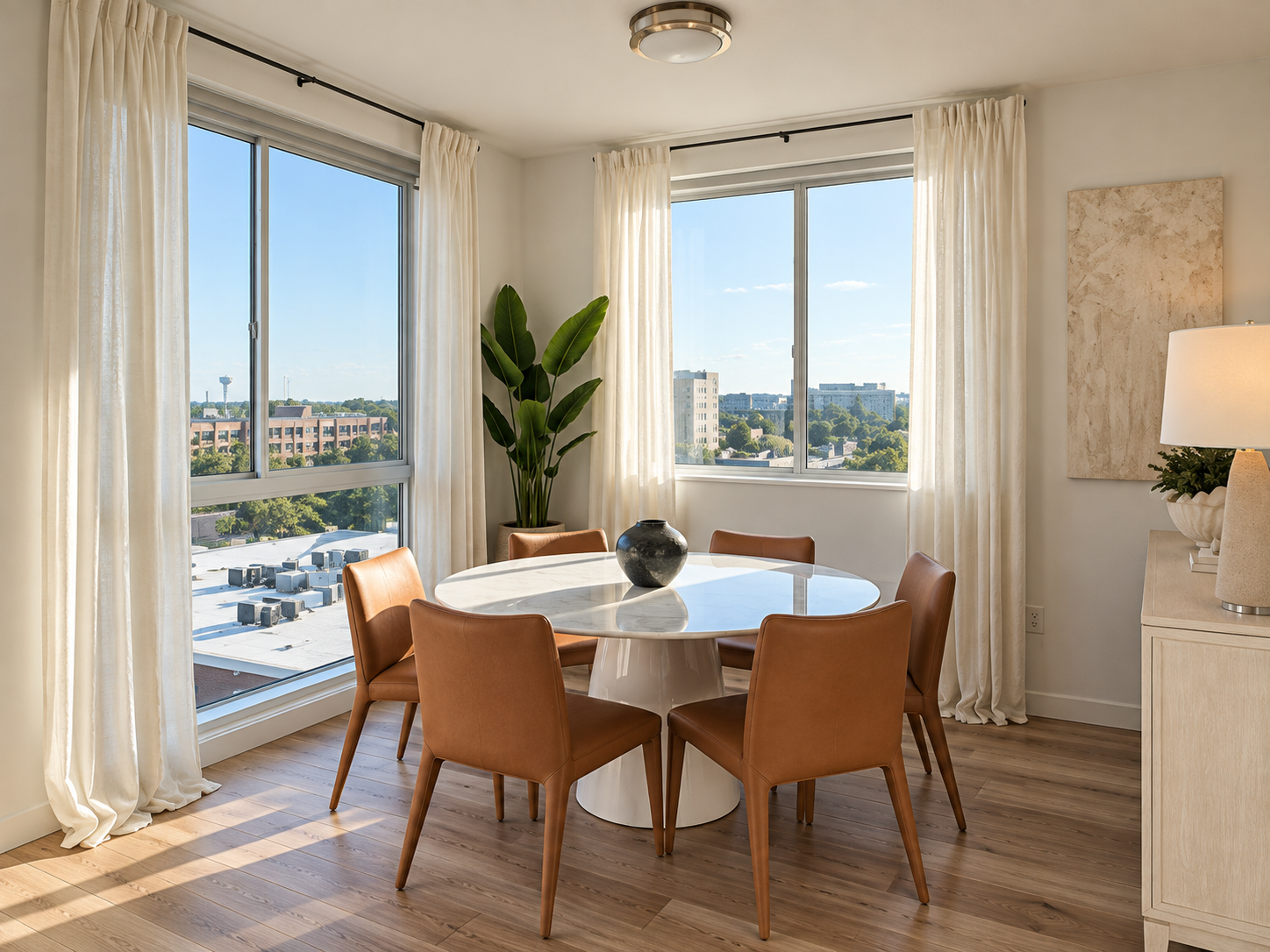 Dining room with a round marble table and six tan leather chairs, large windows with cream curtains, a large green plant, a black vase, a beige cabinet with a lamp, and city view outside.