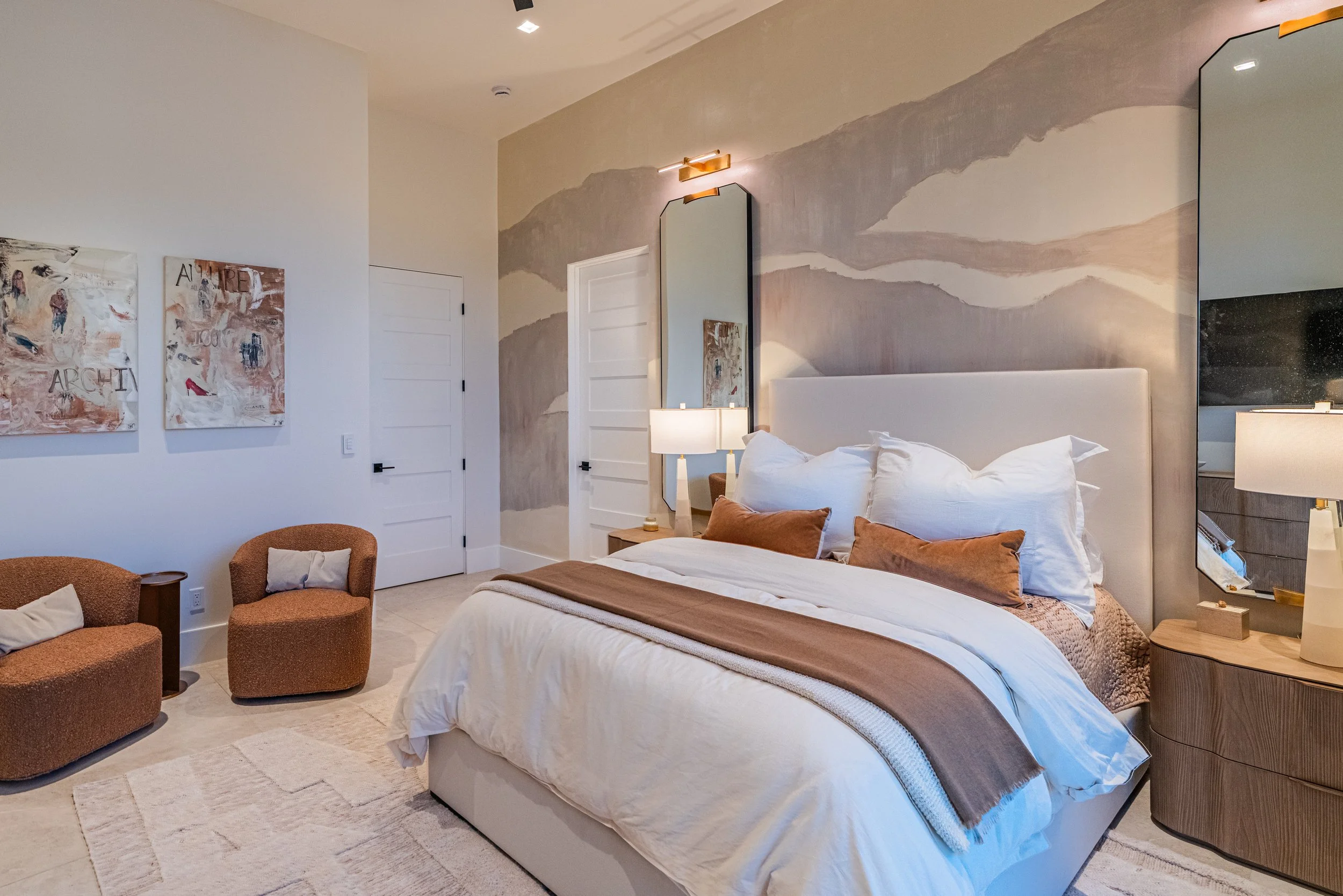 Modern bedroom with a large bed, white bedding, and decorative pillows, flanked by matching bedside tables with lamps, a wall mirror, artwork on the walls, and cozy chairs nearby.