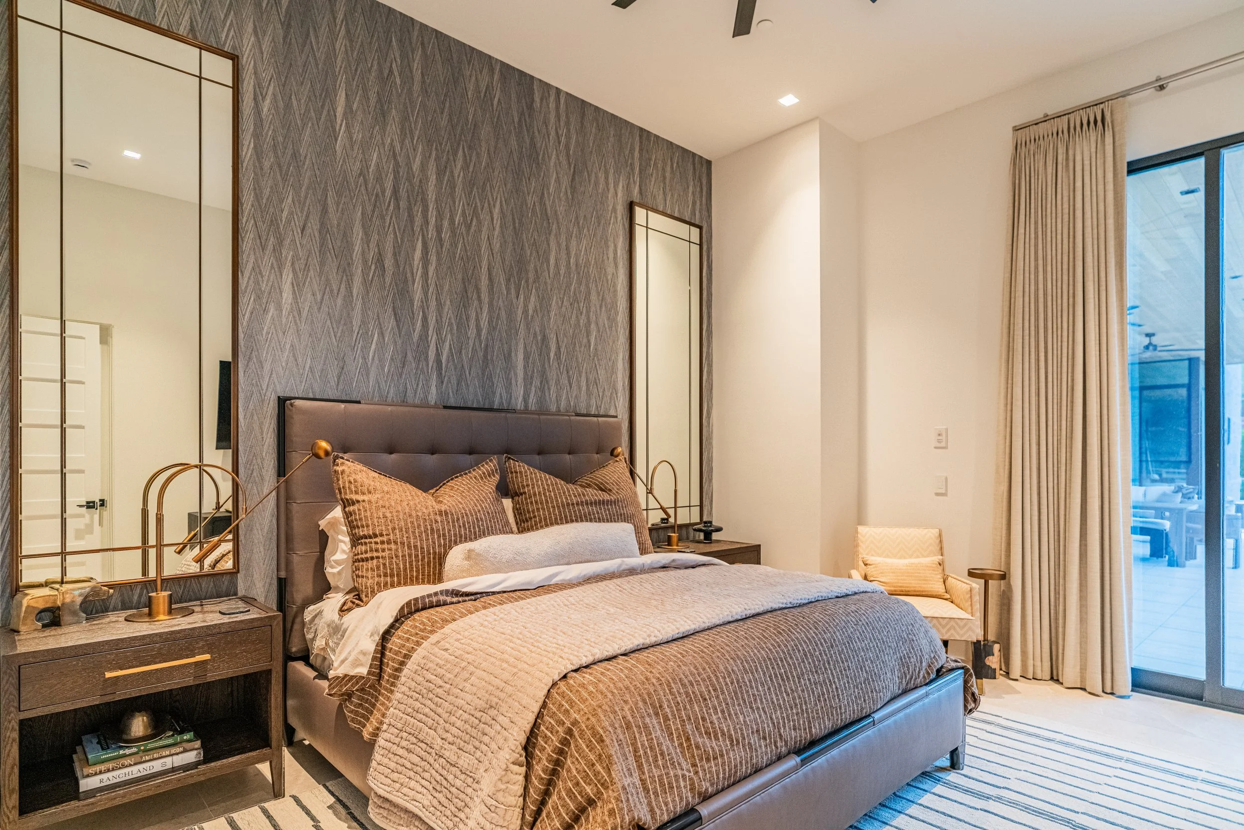 A bedroom with a gray upholstered bed, matching bed linens, and pillows. To the left, there is a wooden nightstand with gold-colored lamps, books, and other decor. There is a large textured mirror above the nightstand. The wall behind the bed has a p