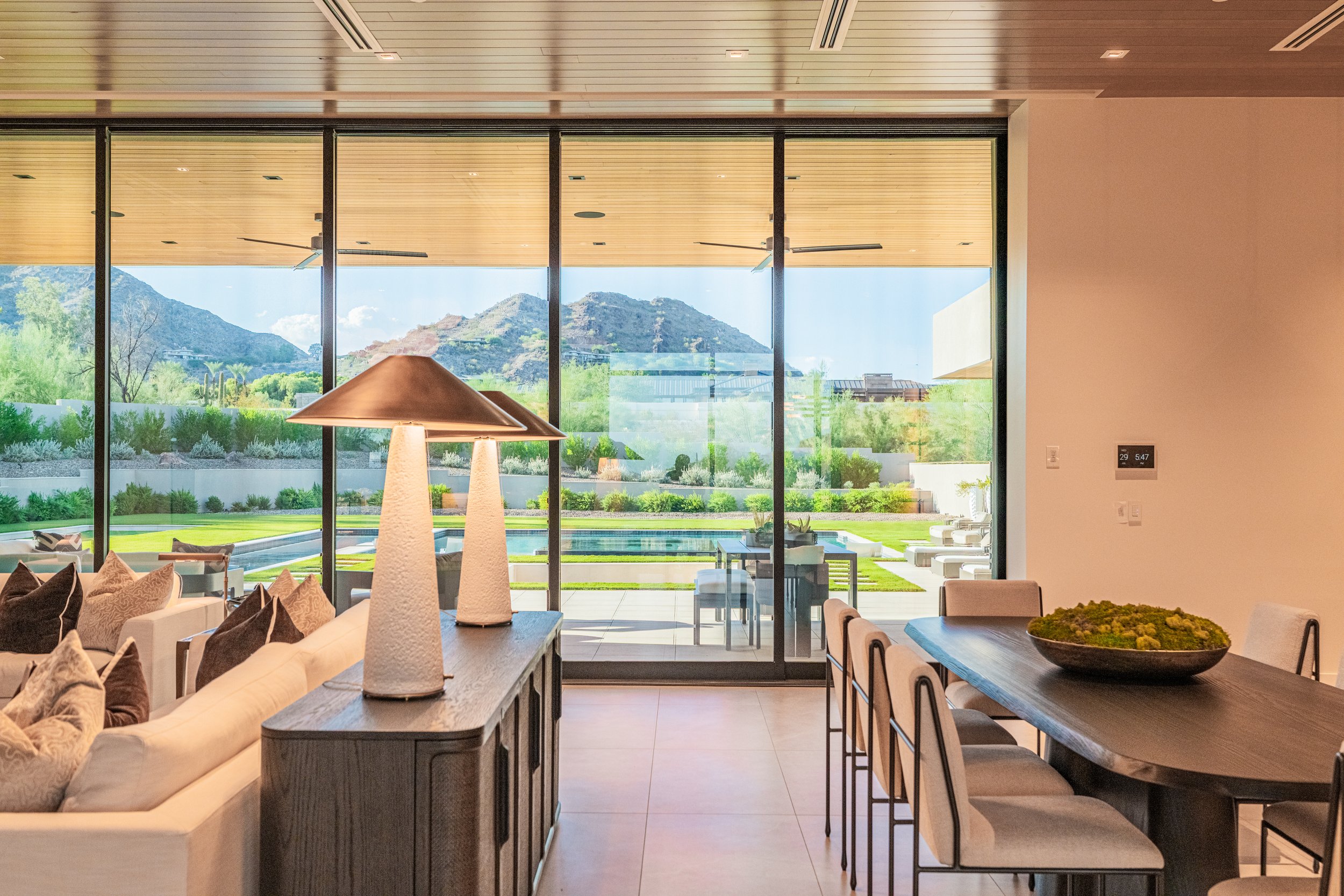 Modern living room with floor-to-ceiling glass windows showing scenic mountains and greenery outside. Inside, there is a long dining table with chairs, a white sofa with pillows, and decorative lamps.