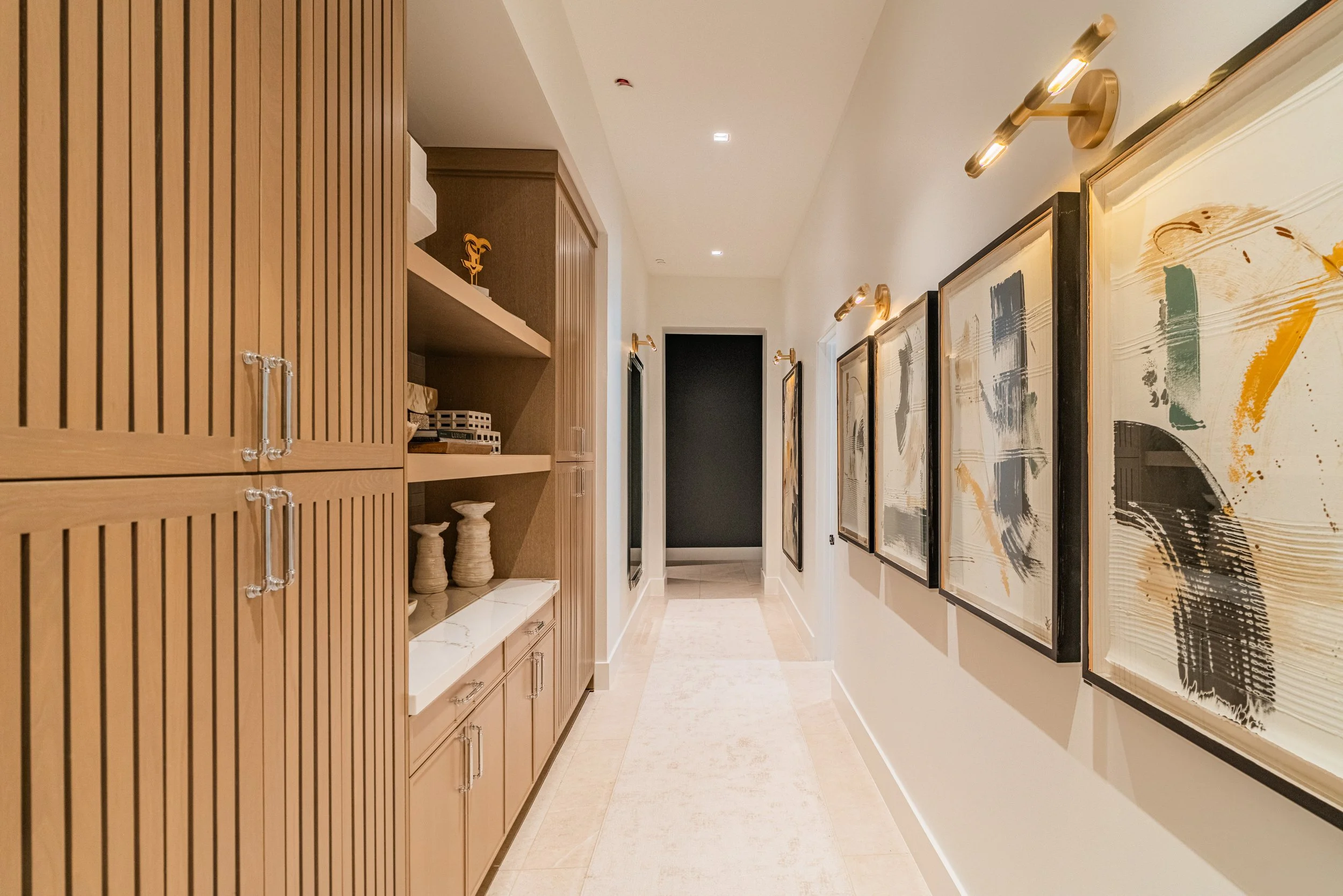 A hallway with multiple framed abstract paintings on the right wall, wooden cabinets and shelves on the left, decorative vases and books on the shelves, warm lighting, and a light-colored tiled floor.
