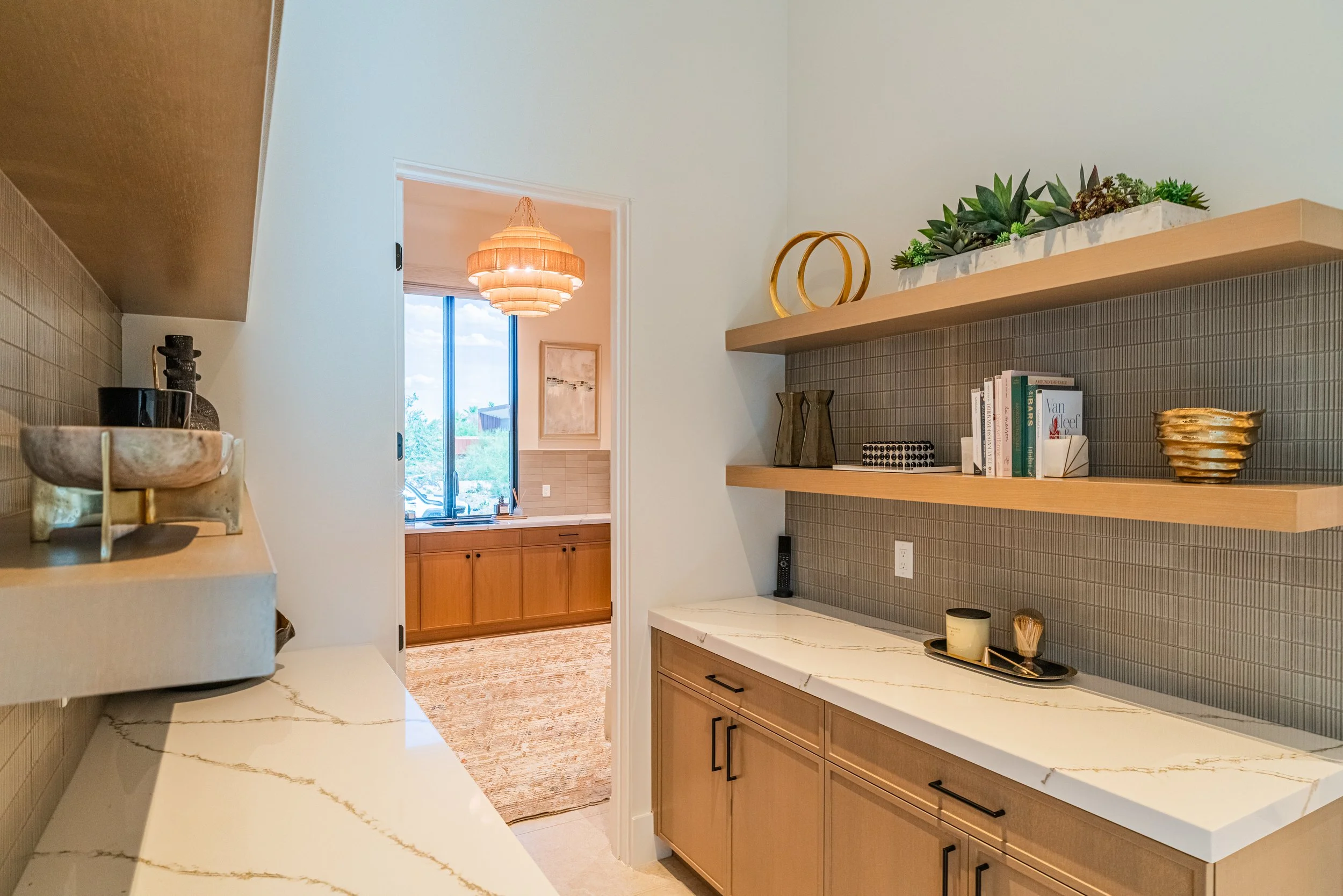 A modern kitchen with beige cabinets, a white marble countertop with gold veining, and a grey tiled backsplash. Decor includes a lazy Susan tray with cups, a vase, and a tray with a candle, in front of a wood shelf holding books, vases, a metallic bo
