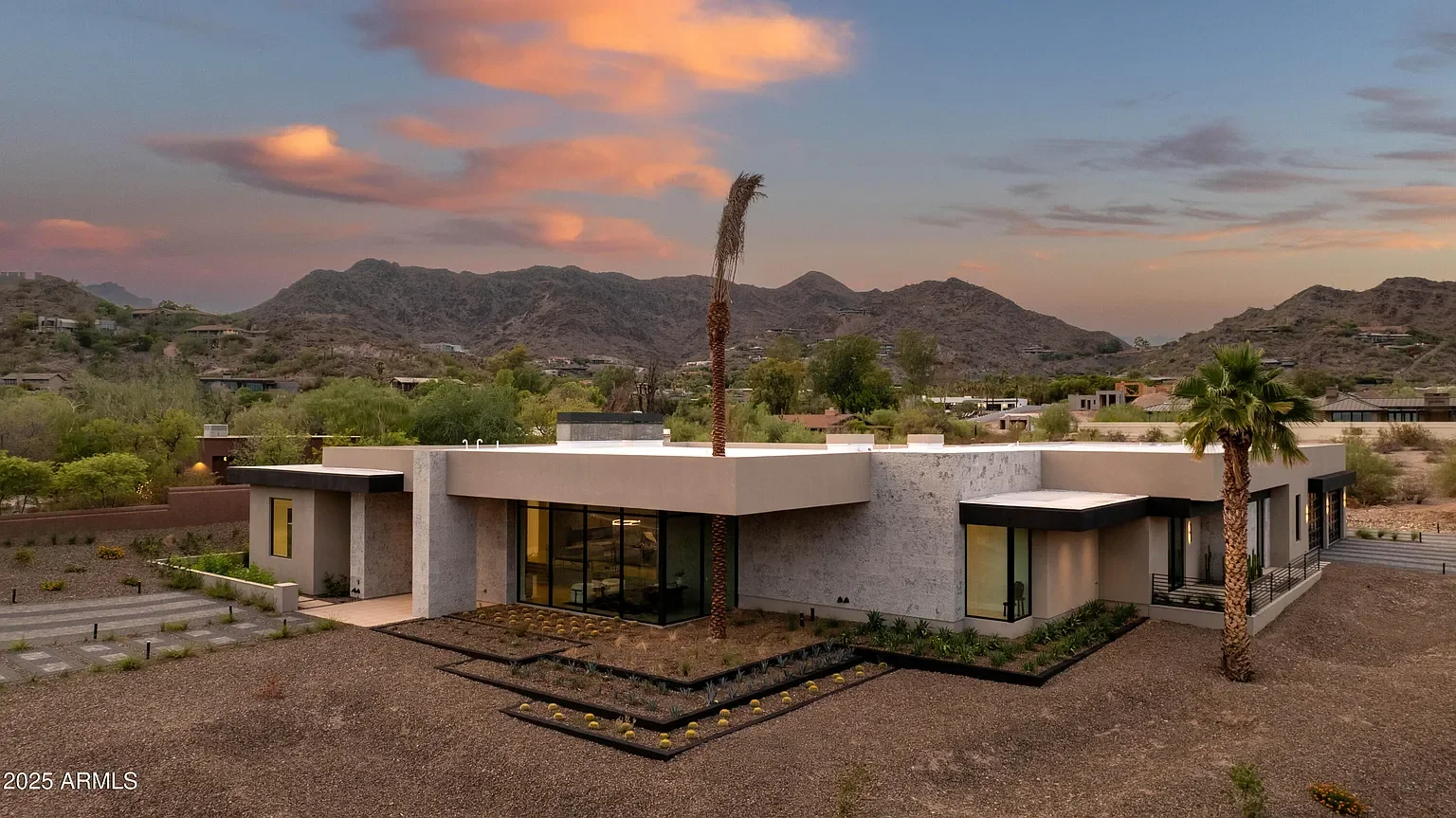 Modern single-story house with large glass windows, surrounded by desert landscape with palm trees, mountains in the background, and a sunset sky.