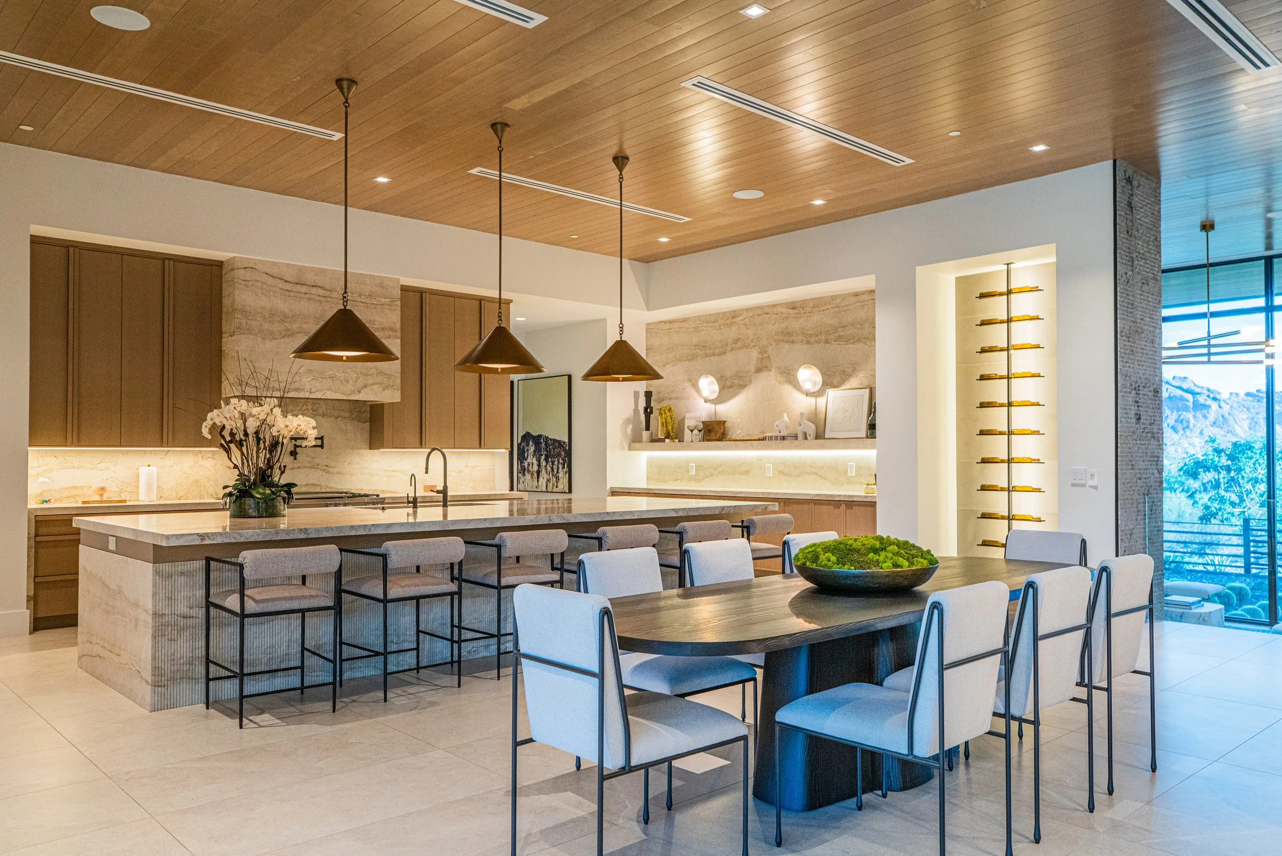 Modern open-concept kitchen and dining area with wood ceiling, pendant lights, large table with white chairs, decorative plant, and large windows showing outdoor scenery.