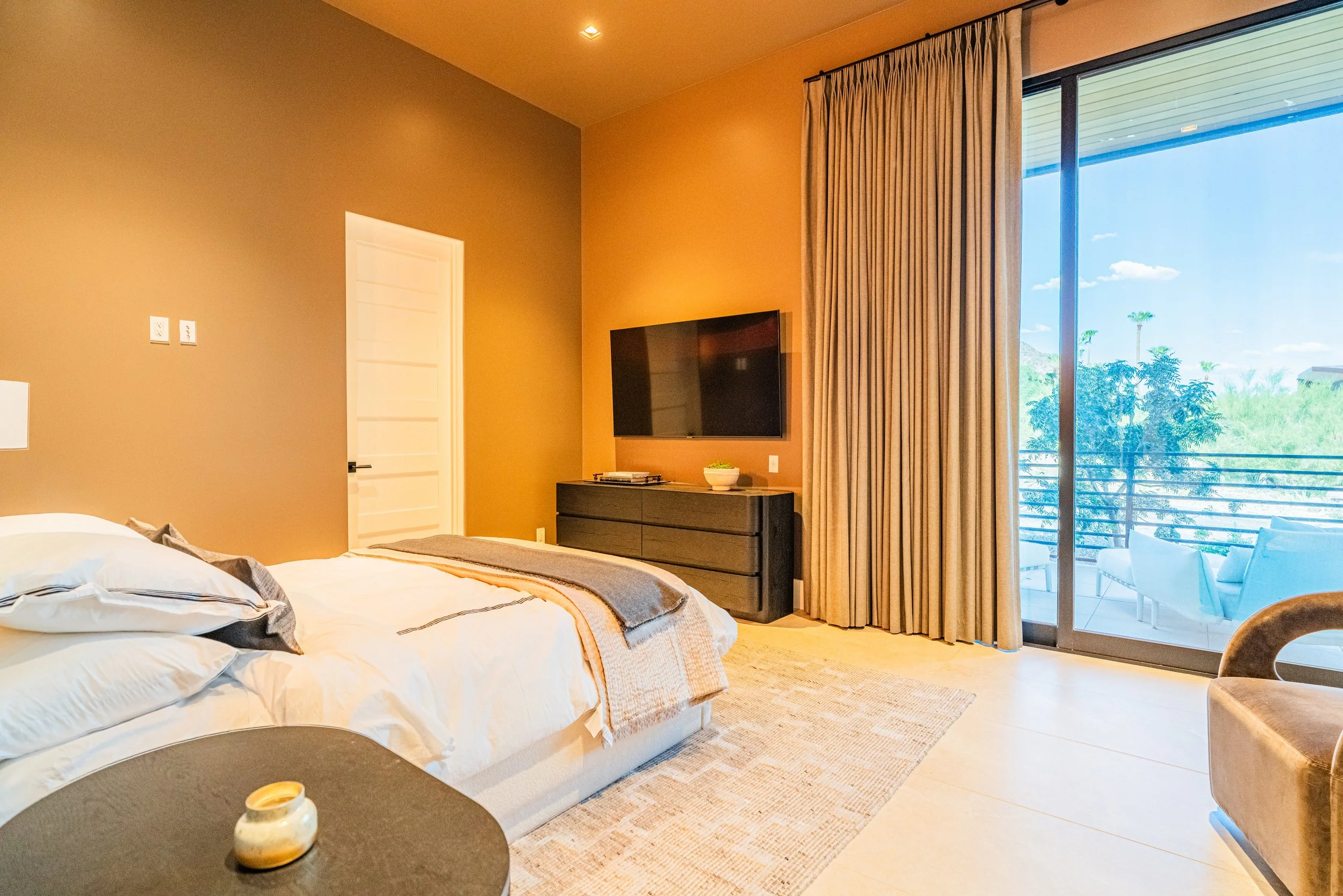 A bedroom with a large bed, a TV mounted on the wall, a black dresser with a bowl on top, beige curtains, and a sliding glass door leading to a balcony with outdoor chairs, overlooking trees and a clear blue sky.