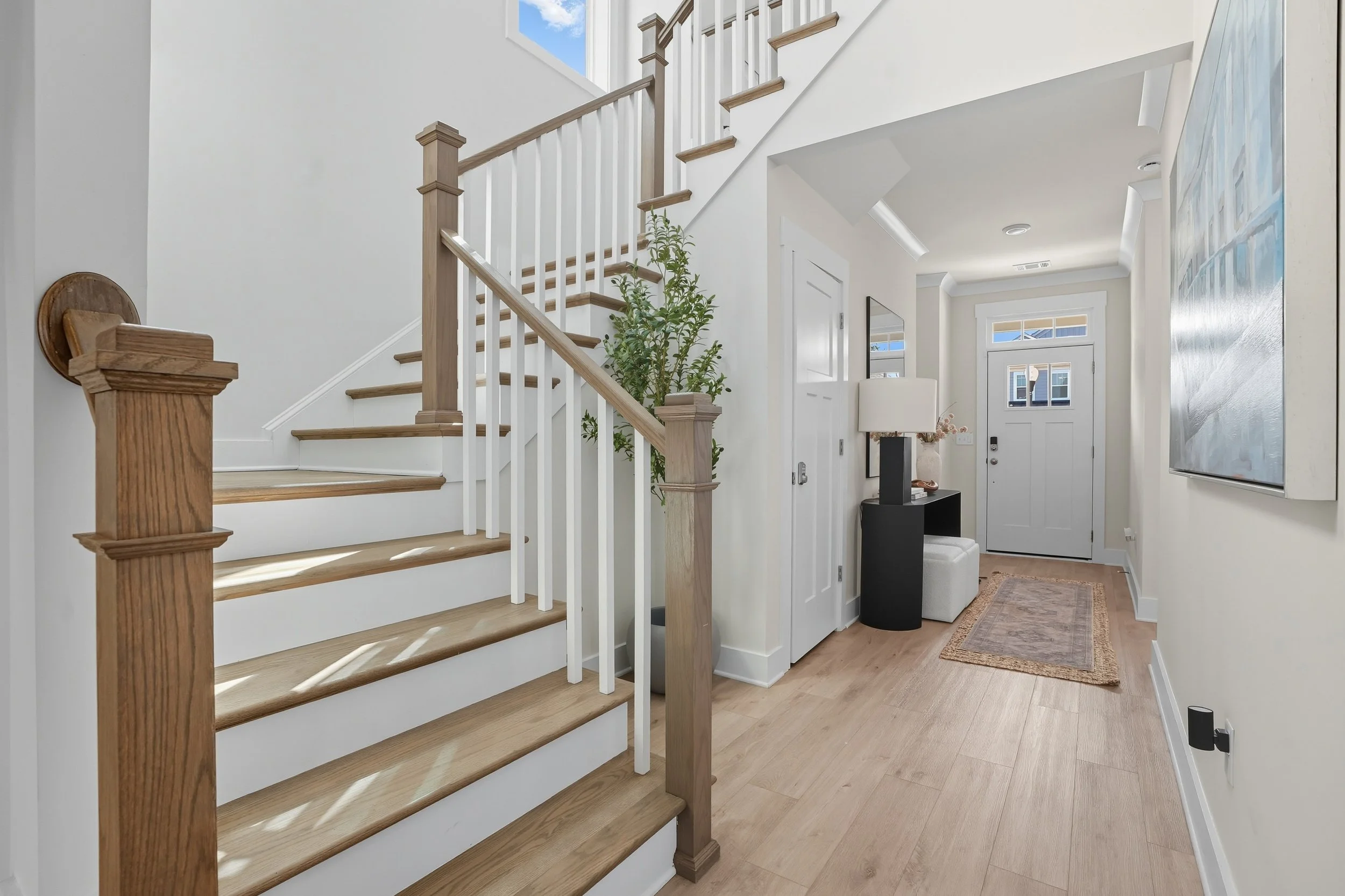 Interior view of a home with a wooden staircase, white walls, and a front door with windows. There is a table with a lamp near the entrance, a carpet on the floor, and a tall potted plant.