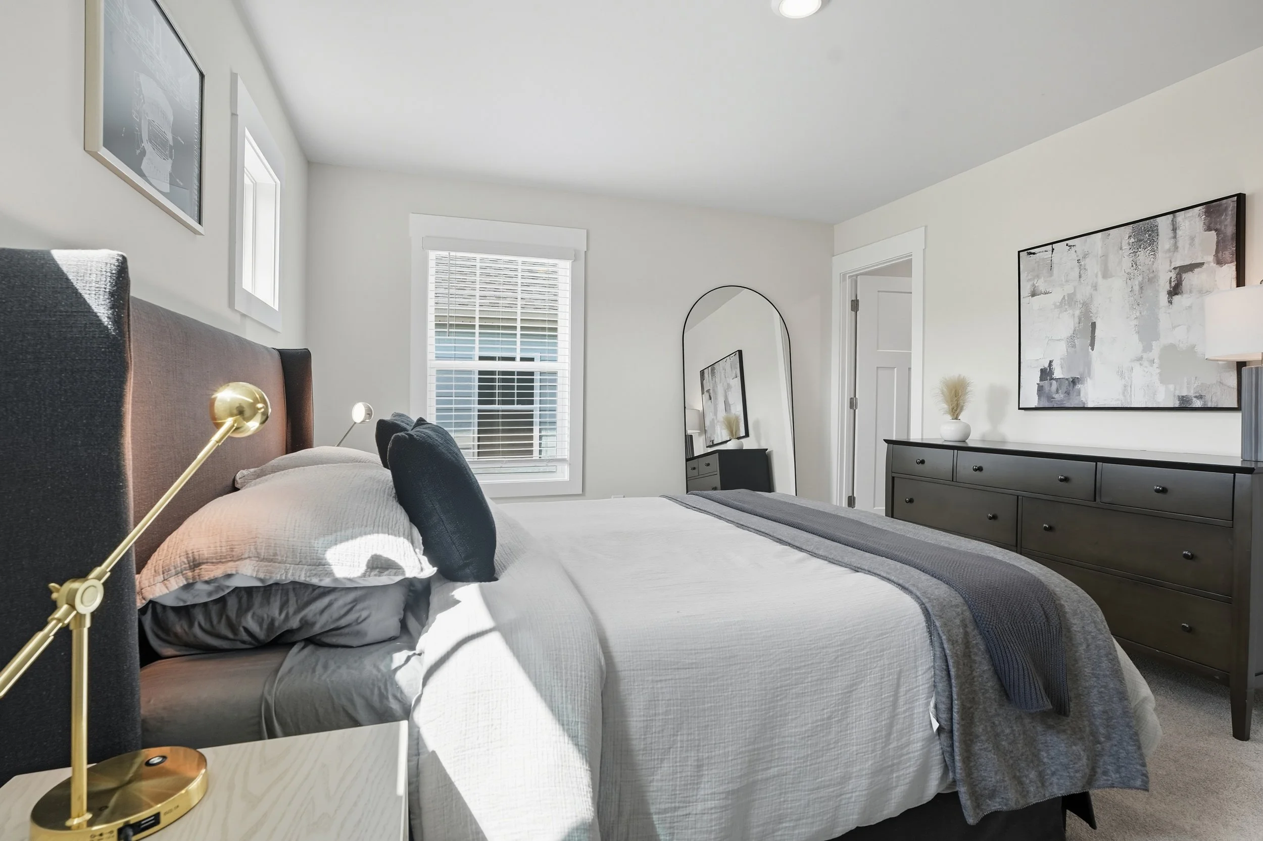 A modern bedroom with a bed that has a brown upholstered headboard, gray bedding, and black accent pillows. Beside the bed is a white nightstand with a gold lamp. The room has two windows with white blinds, white walls, and contains a large black dre