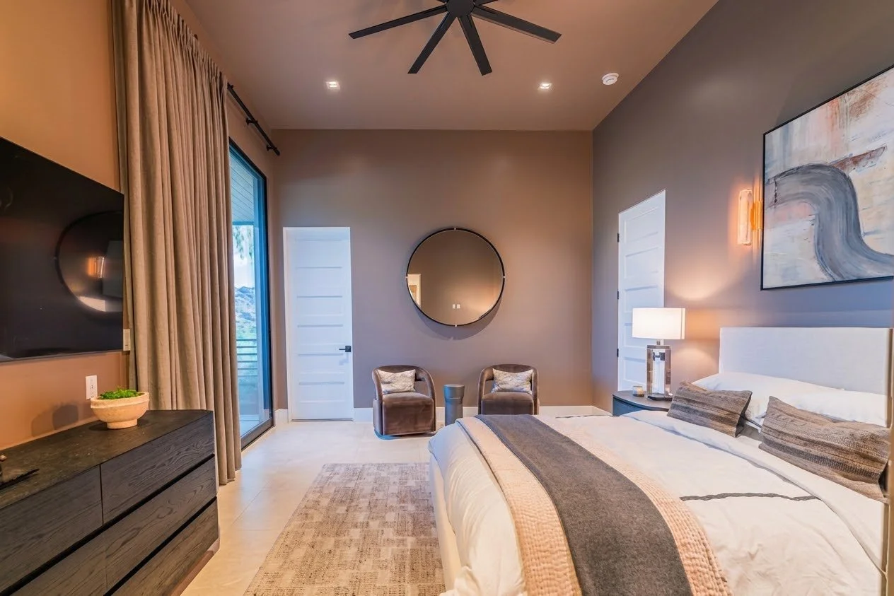 Modern bedroom with gray walls, large bed with white bedding and pillows, black dresser with TV, sliding glass door with curtains, round mirror on wall, two brown chairs with cushions, bedside table with lamp, abstract painting on wall.