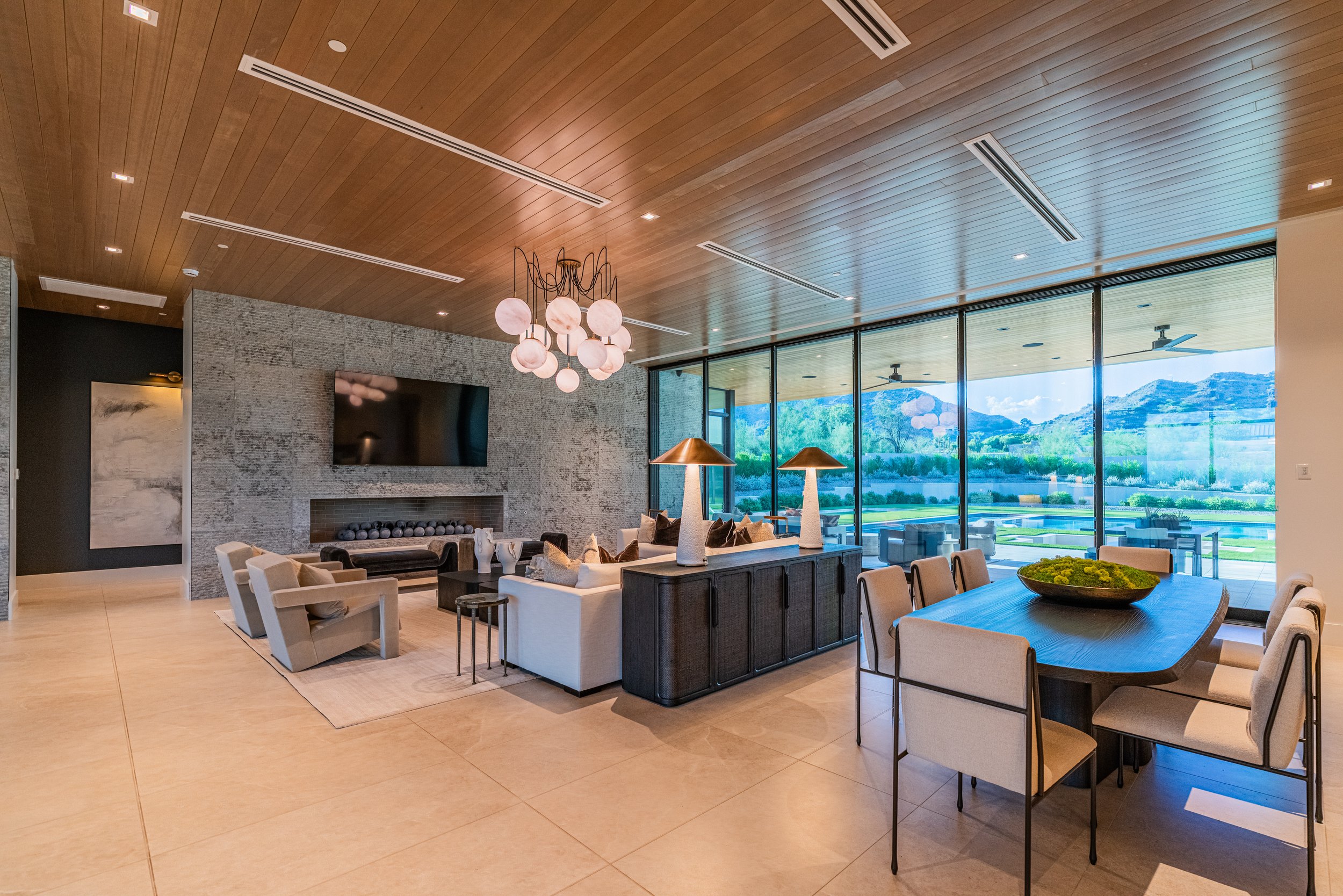 Bright and modern living room with a wooden ceiling, large windows showing a scenic outdoor view of mountains and greenery, a stone fireplace, a chandelier with multiple globes, a long dining table with beige chairs, and decorative lamps.