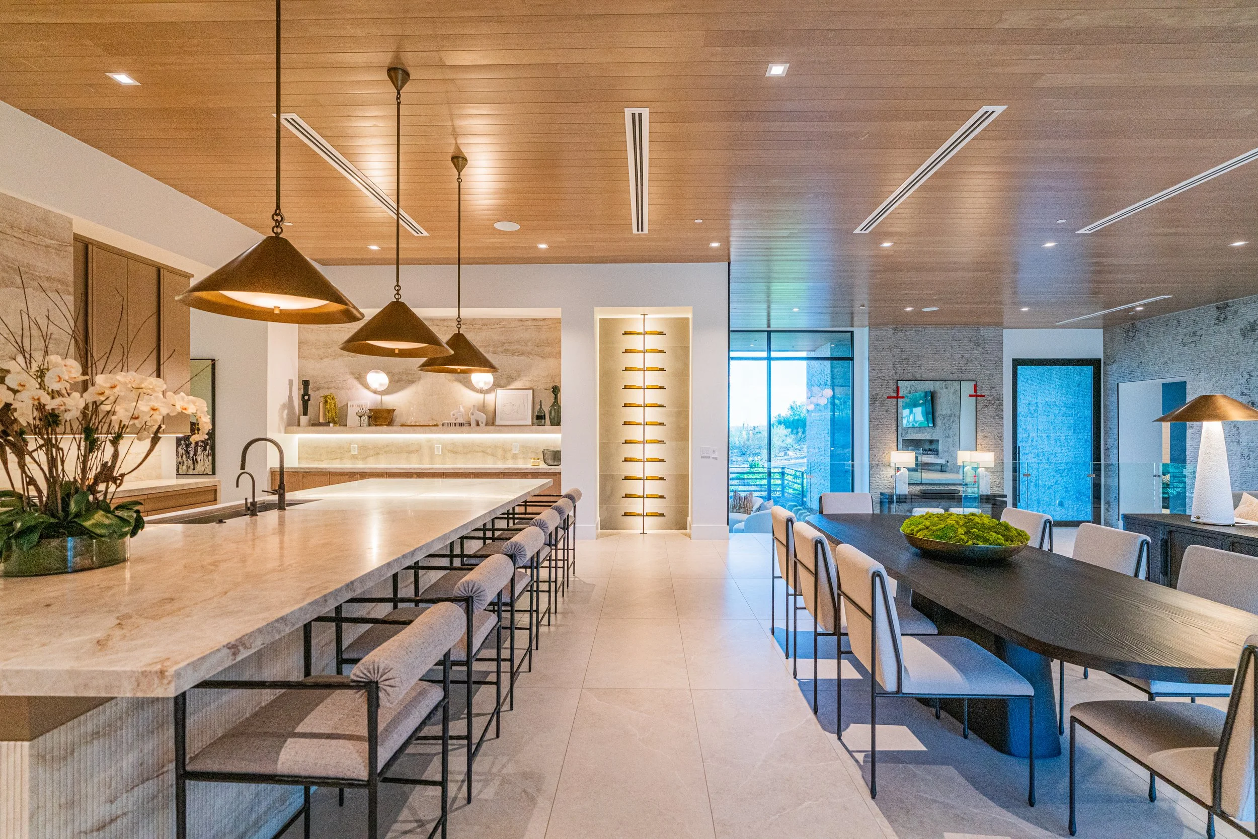 Modern open-plan kitchen and dining area with a large marble island, pendant lights, a wooden ceiling, and a glass door leading outside.