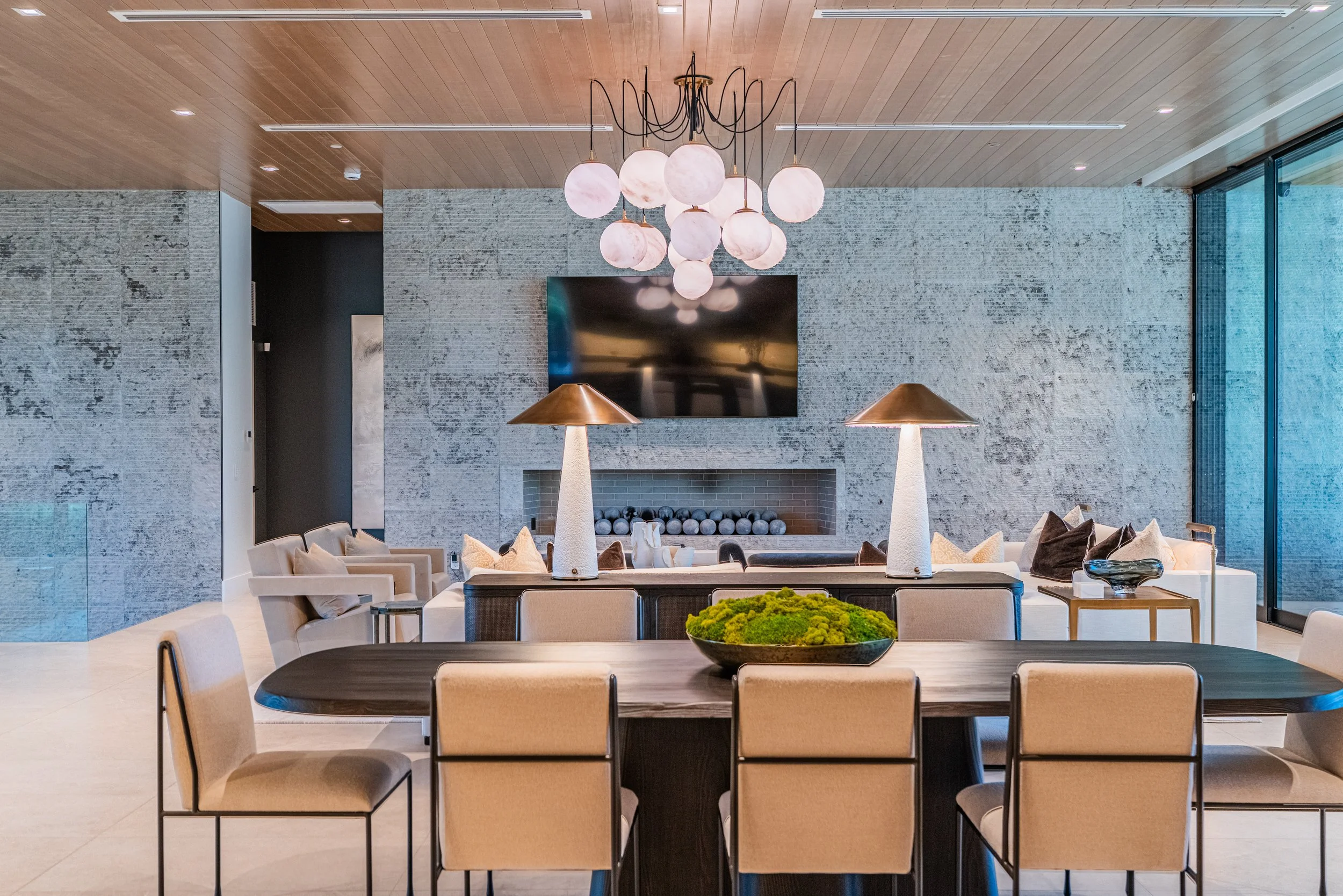 Modern living room with a wooden ceiling, abstract wall art, large flat-screen TV, pink glass globe chandelier, cream chairs, and a black dining table with beige chairs, decorative lamps, and a centerpiece.