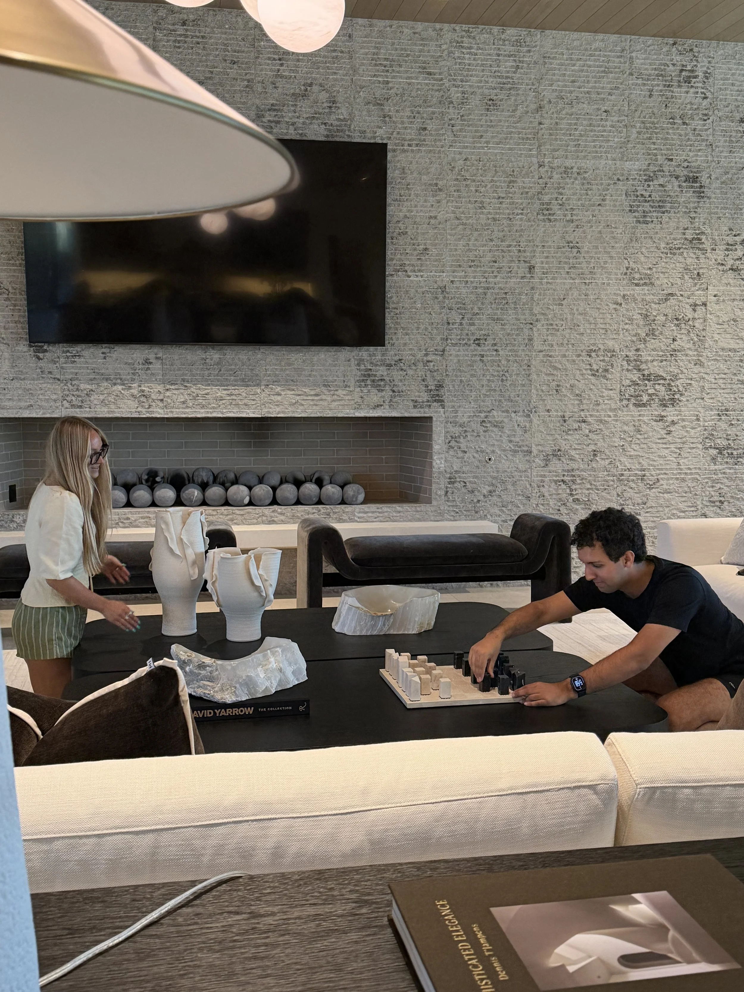 People in an interior space with a large textured gray wall, a black television, and a decorative fireplace with spherical objects. Two individuals are at a table with white and black sculptures and a chess game. One person with long blonde hair and glasses is standing, and another person with dark hair is seated, reaching towards the chess pieces.