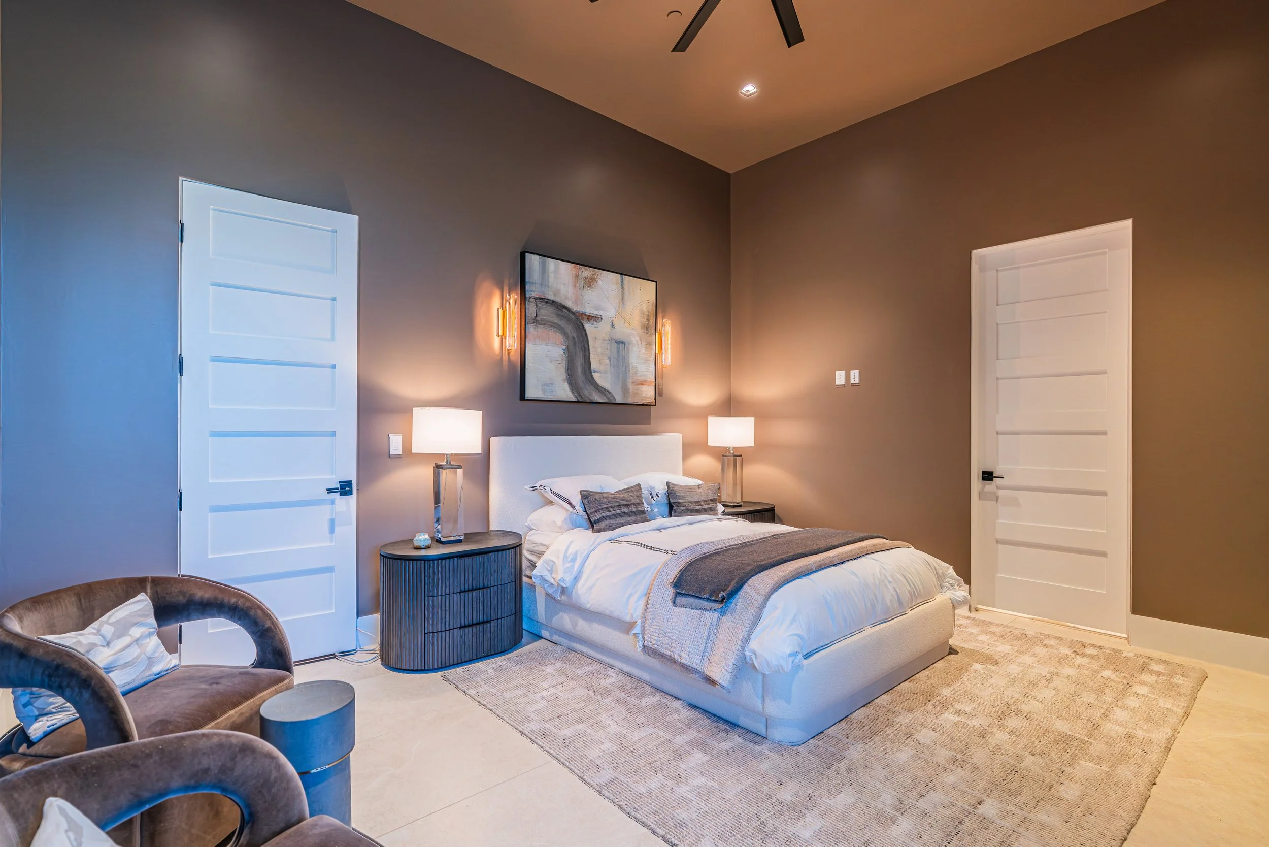 A modern bedroom with a bed, two matching nightstands with lamps, and abstract artwork on the wall. The room has beige walls, two white paneled doors, and a ceiling fan. There's a sitting area with two brown velvet chairs and a small round table.