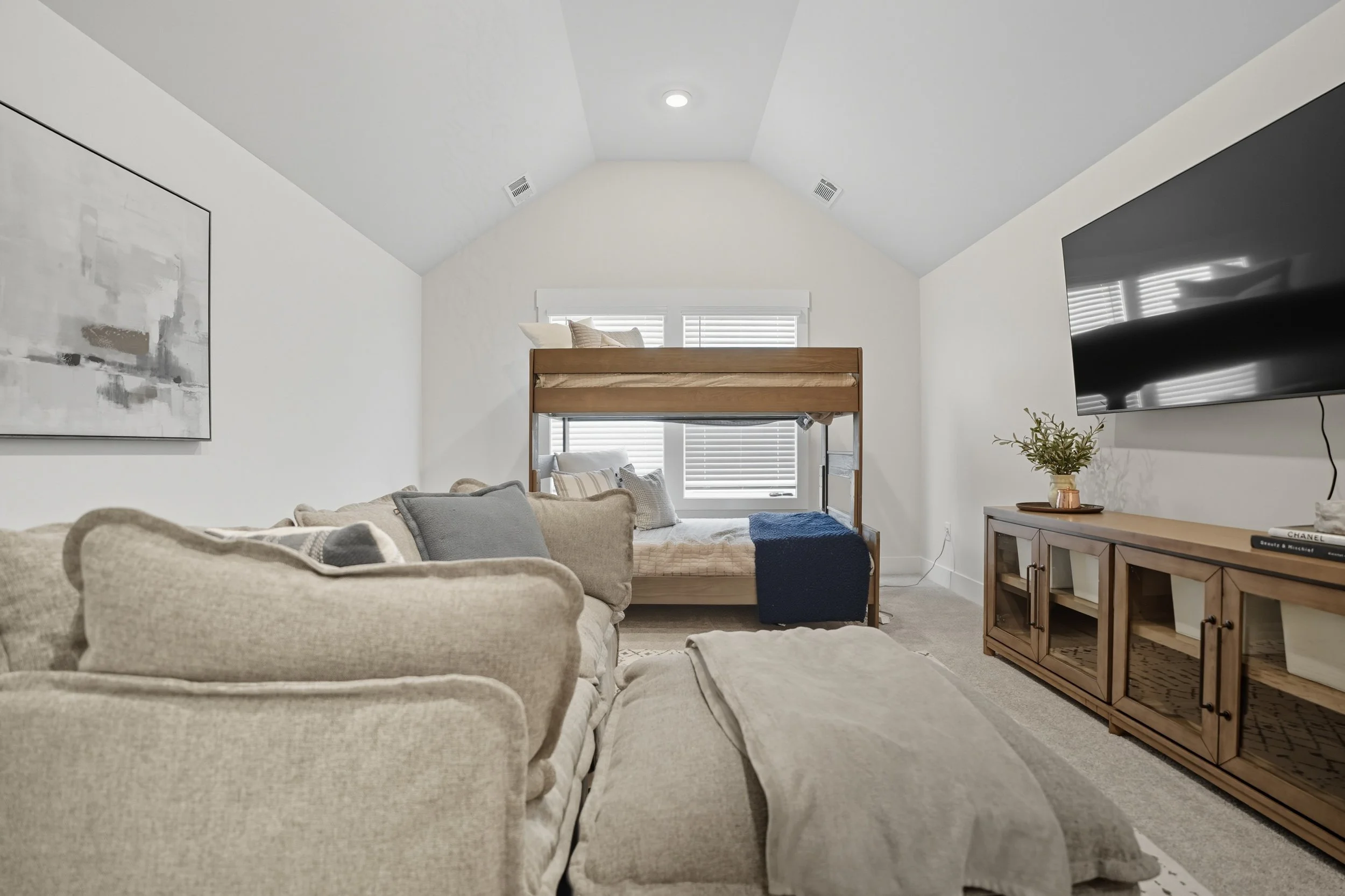 A cozy living room with a beige sofa, a wooden bunk bed near a window with blinds, a large wall-mounted TV, and a wooden cabinet with decorative items, including a small plant and books.
