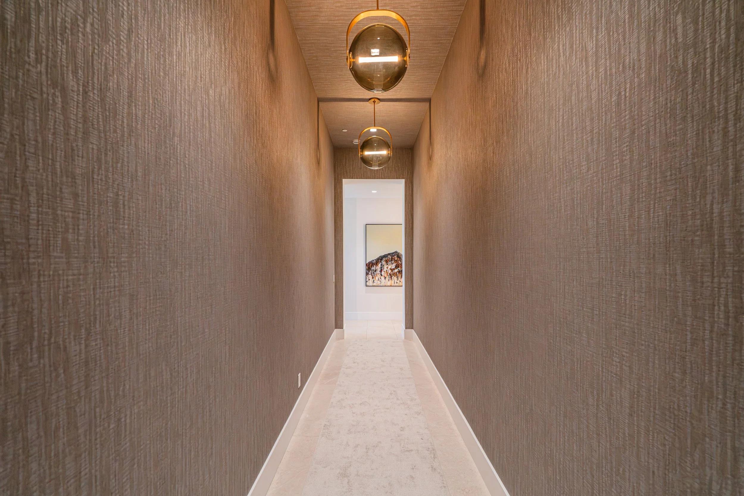 A long hallway with textured beige walls, a light-colored tiled floor, and hanging oval-shaped pendant lights with golden accents. There's a doorway at the end of the hallway with a painting visible inside.