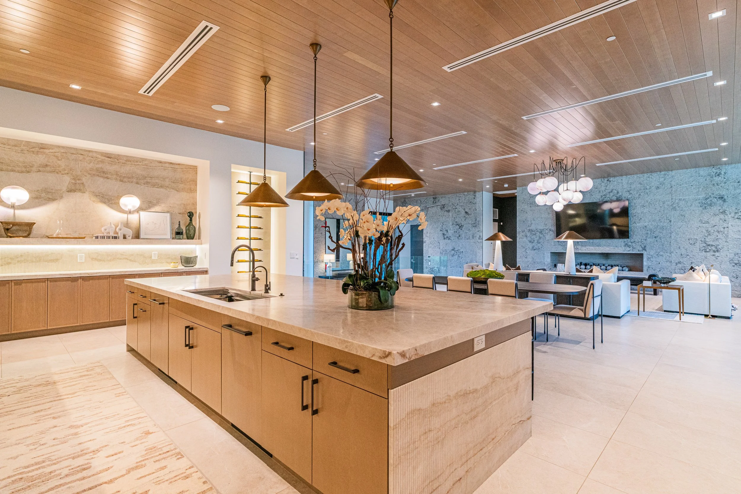 Modern kitchen and living room with wooden ceiling, pendant lights, large island with flower vase, and a wall-mounted TV.