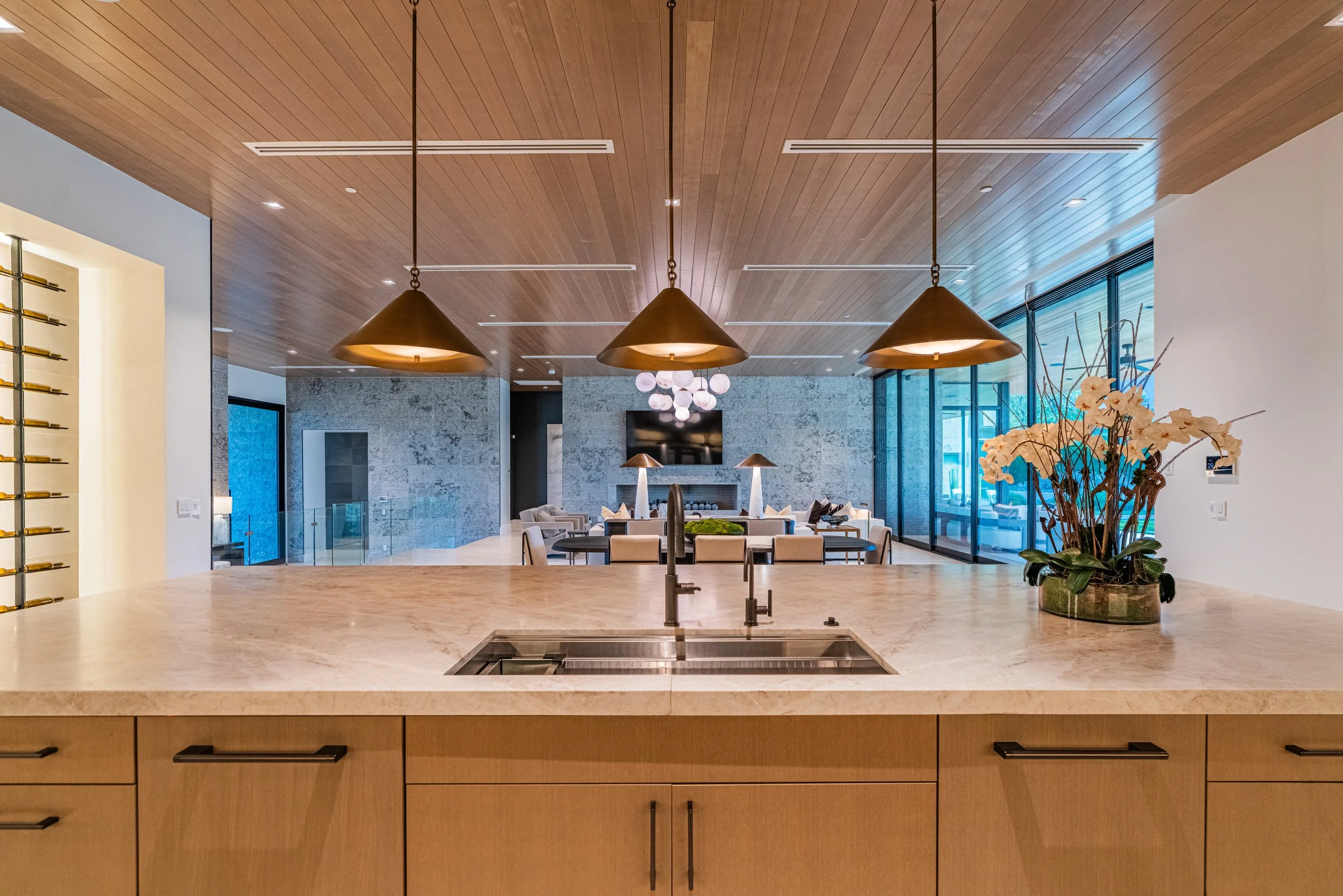 Modern kitchen with a beige marble island, suspended copper pendant lights, and a view of a spacious living area with large windows, a television, and decorative elements like a plant with white flowers.
