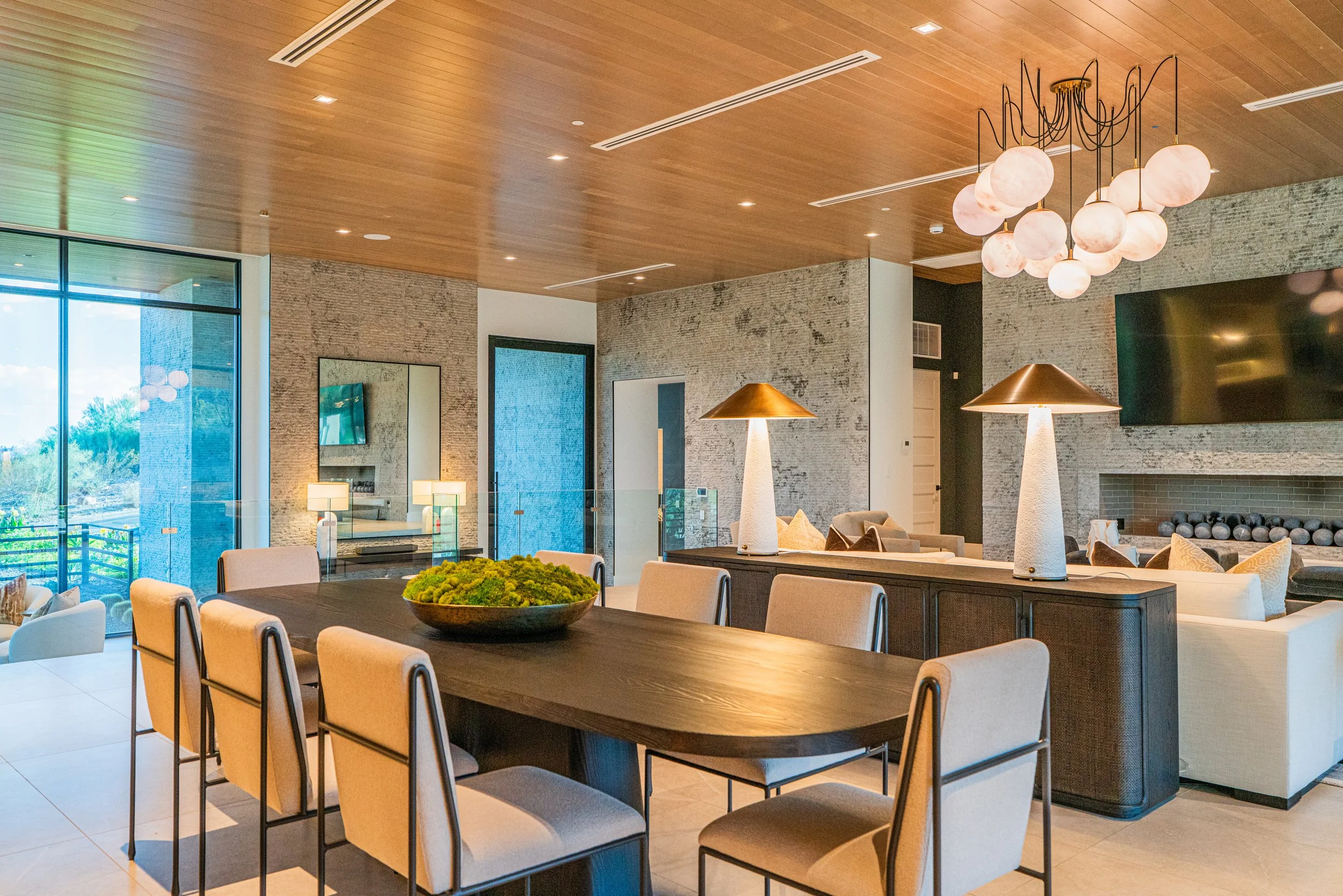 Modern dining and living area with wooden ceiling, stone walls, and large windows. Includes a dining table with chairs, decorative lamps, chandelier, and a fireplace.