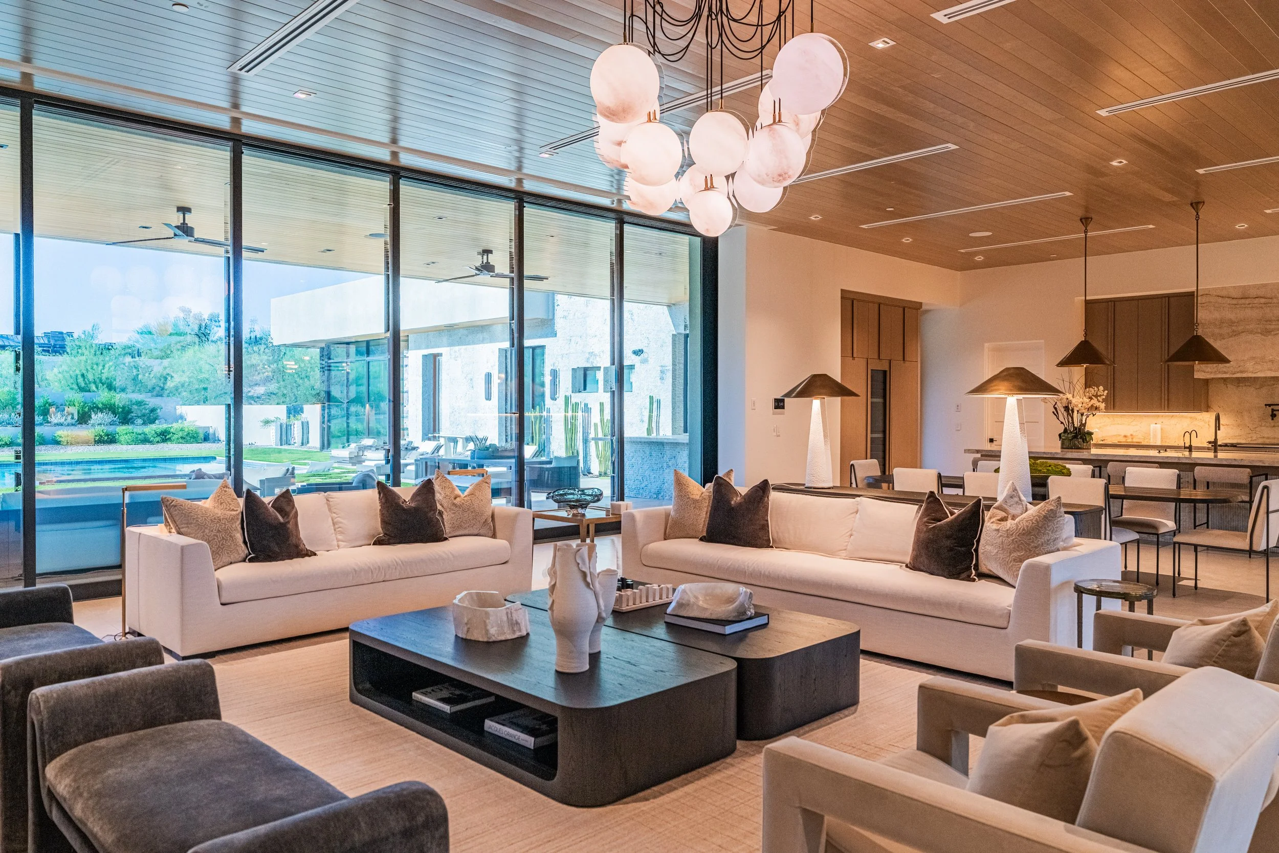 Modern living room with large windows, cushioned sofas, and a contemporary chandelier hanging from wooden ceiling.