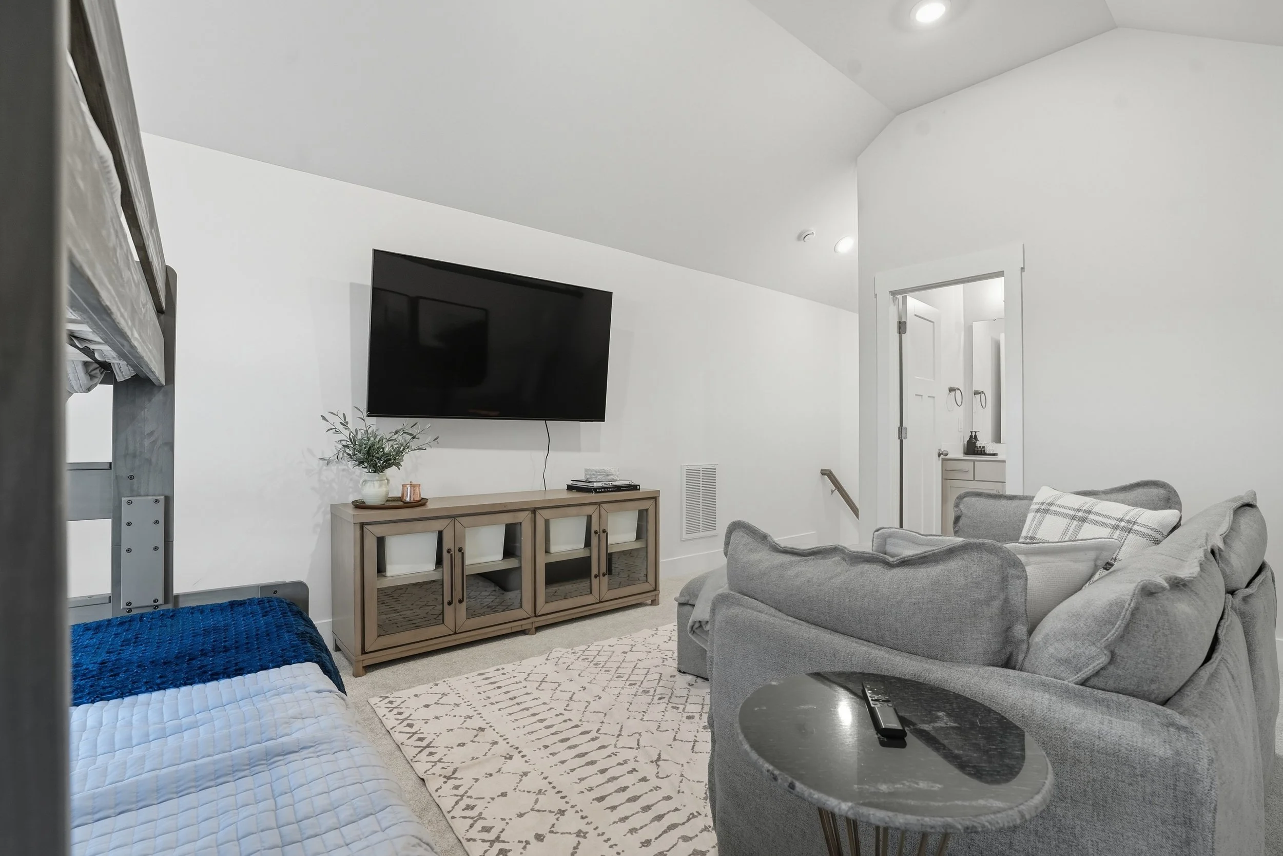 Living room with a mounted flat-screen TV, gray sofa with plaid pillow, wooden storage console with white baskets, a plant, and a rocking chair with a black object on a round side table. White walls and an open door leading to a bathroom.