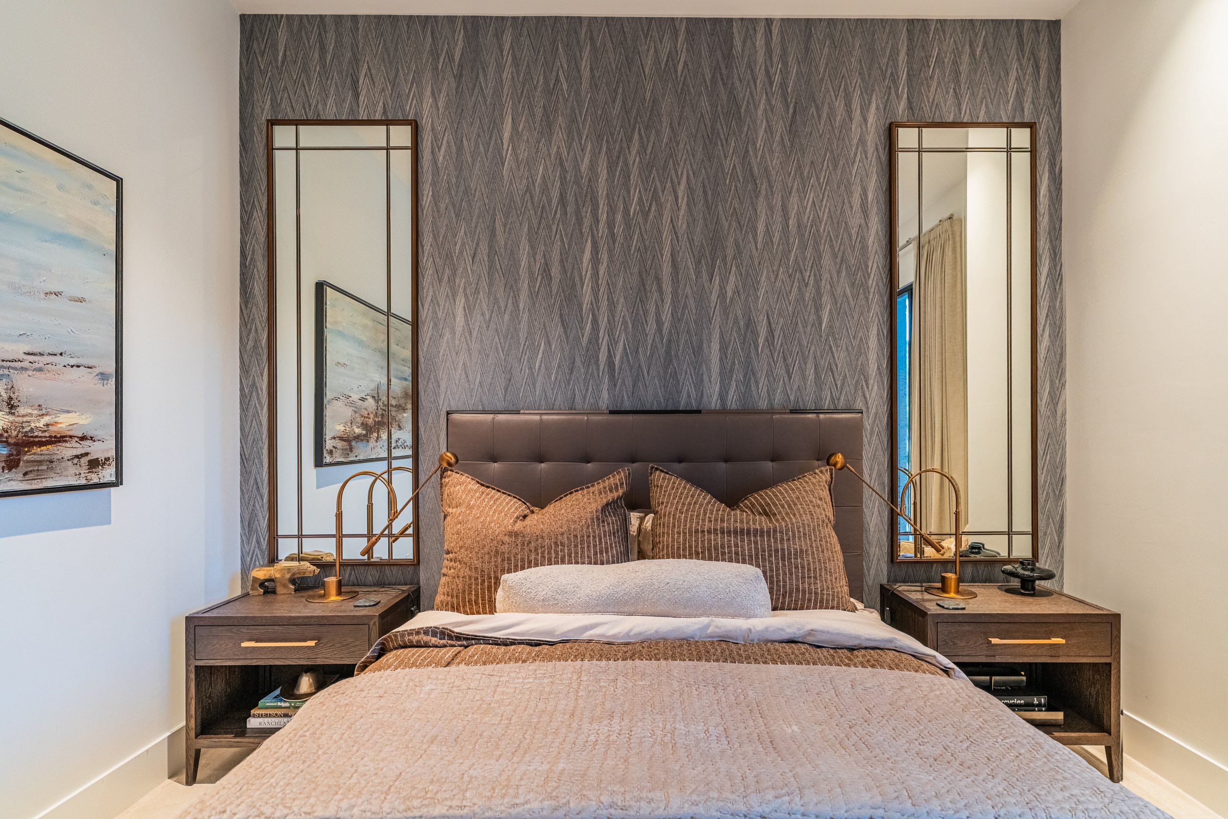 A modern bedroom with a tufted headboard, decorative pillows, and a textured beige bedspread. Two matching wooden nightstands with books, lamps, and decorative items flank the bed. Behind the headboard is a large, patterned gray accent wall with two 