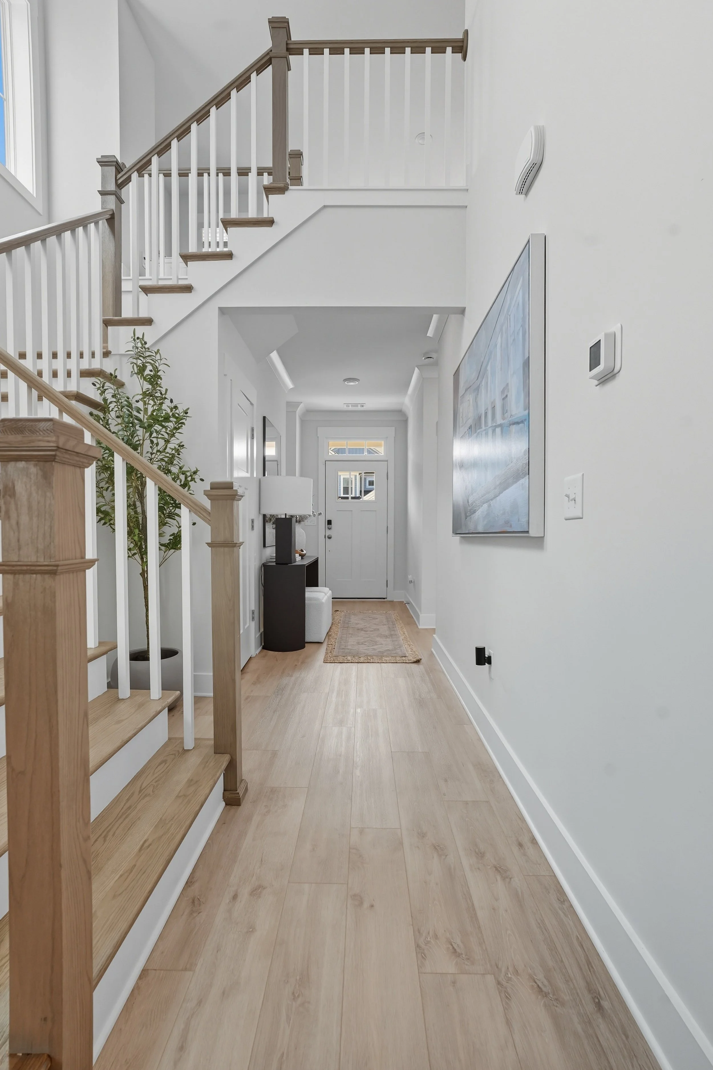 Interior view of a bright, modern home hallway with a staircase on the left, a picture gallery on the right wall, and a front door at the end, featuring light wooden flooring and white walls.