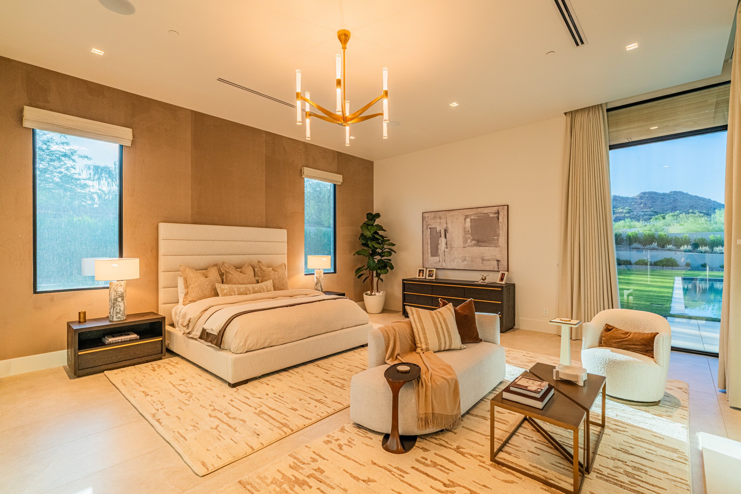 Modern bedroom with beige walls and large windows showing outdoor greenery and mountains. The room has a white bed with beige pillows, a black nightstand with a marble lamp, a potted plant, and a black dresser with artwork. There is a beige rug and w