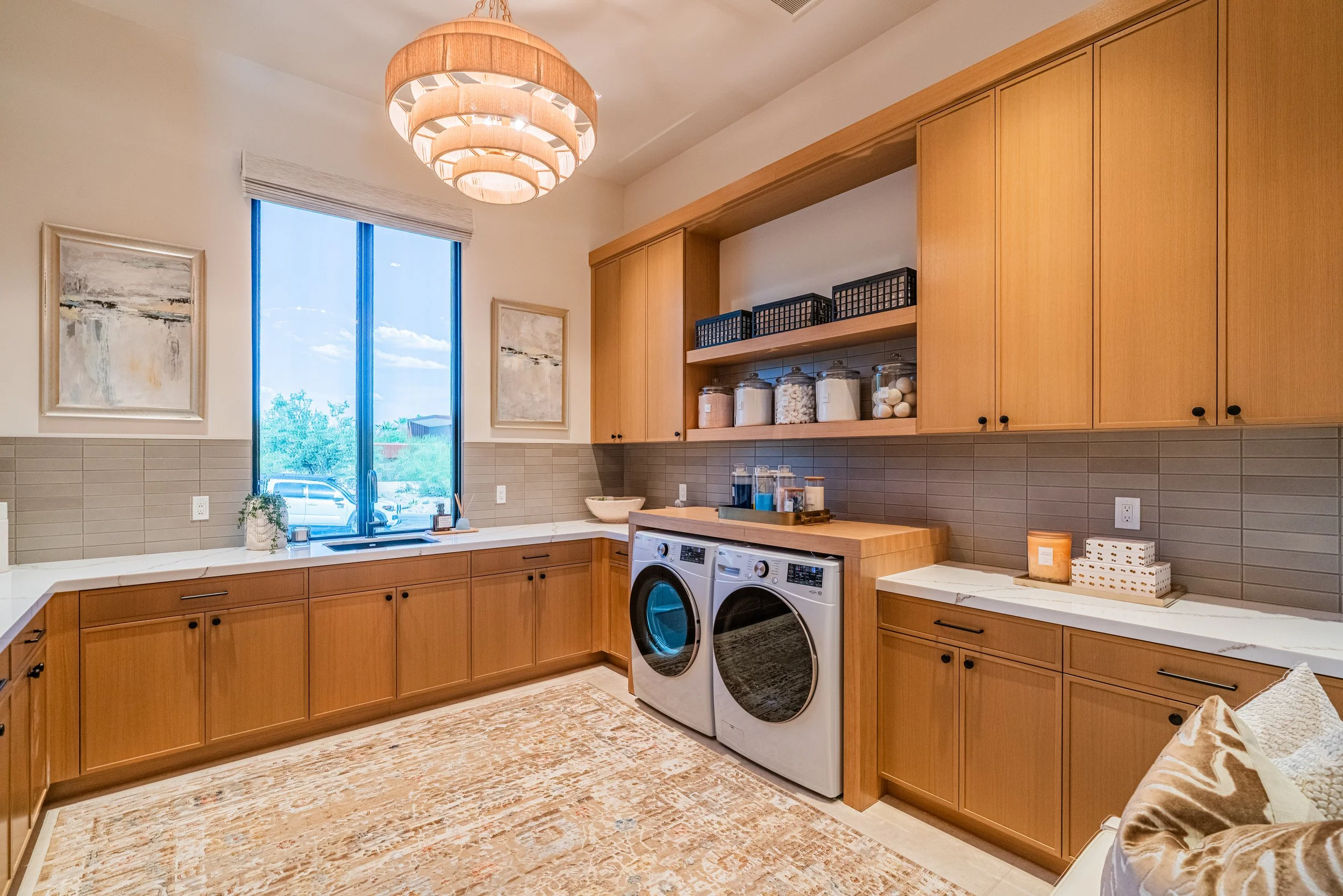 A modern kitchen with light wood cabinetry, a white countertop, and a large window. There are two abstract paintings on the wall, a round wooden chandelier hanging from the ceiling, and a washer and dryer beneath the counter. Open shelves hold contai