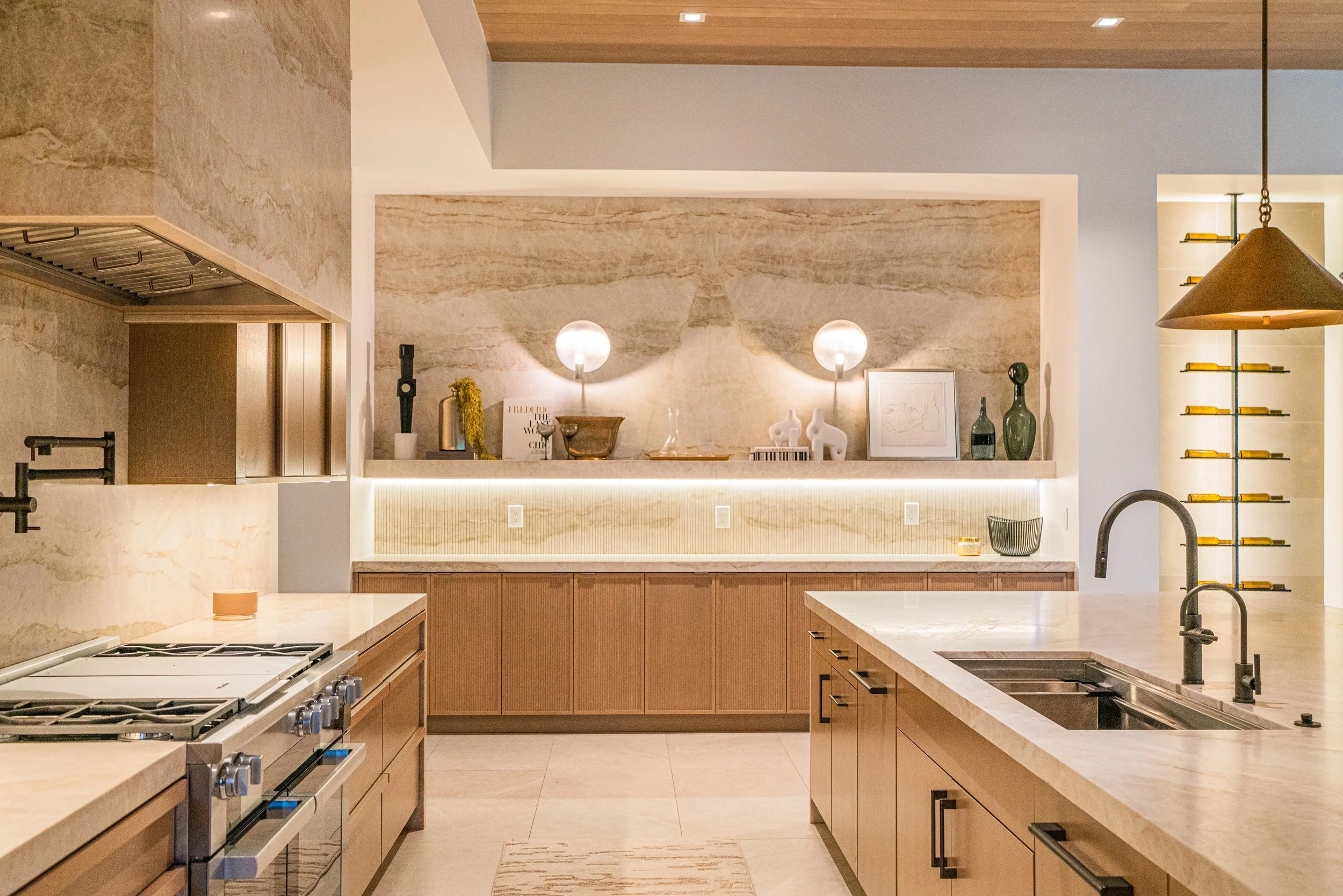 Modern kitchen with beige countertops and wooden cabinets, featuring a stove, under-cabinet lighting, decorative items on a shelf, and a pendant light hanging from the ceiling.
