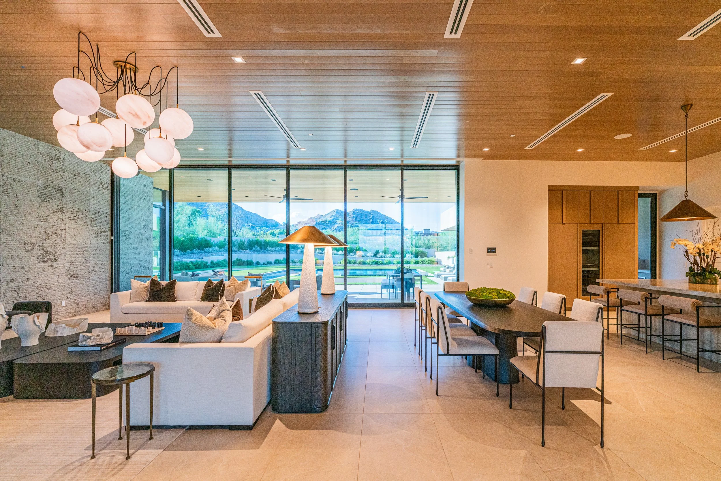 Modern living room with wooden ceiling, large window showing mountains, white sofa with pillows, rectangular dining table with white chairs, decorative lamps, vase of orchids, and outdoor area with pool and garden.