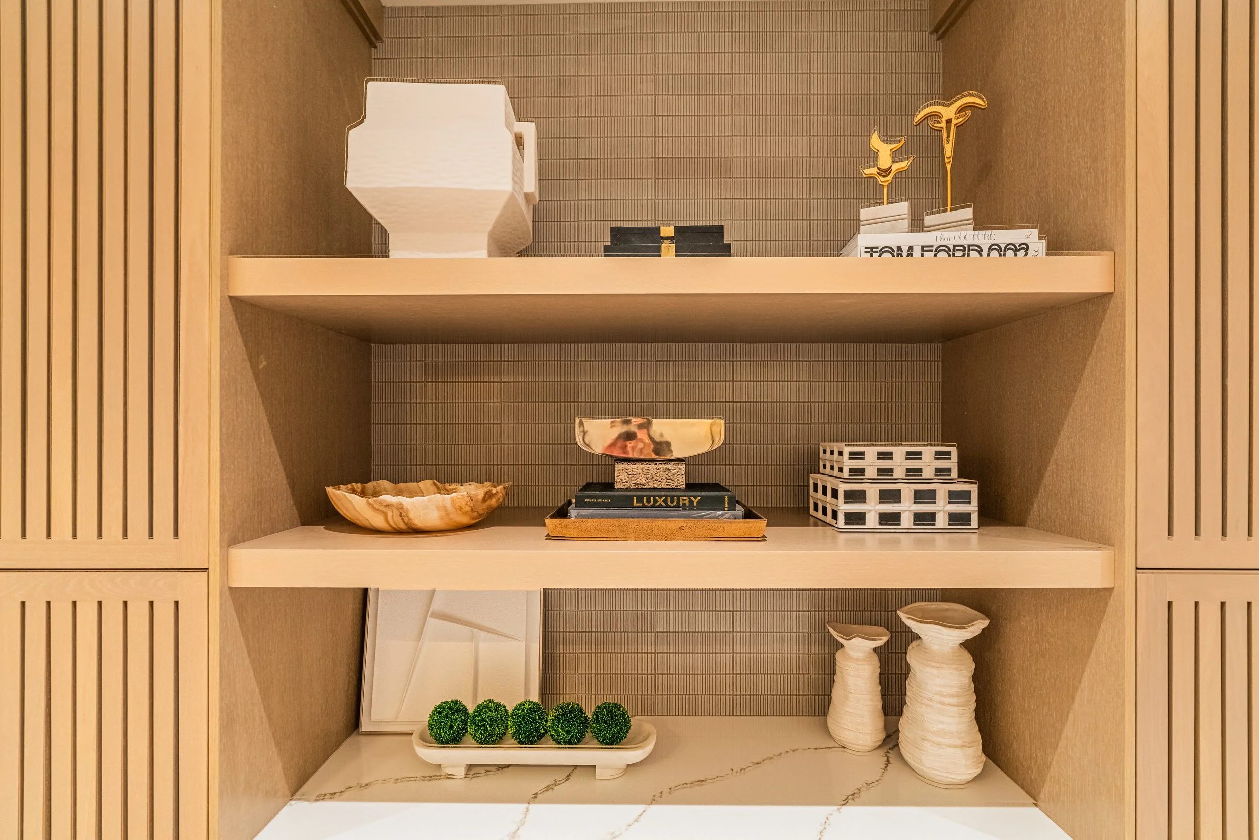 Open beige-colored cabinet with three shelves displaying decorative items such as a white geometric sculpture, black boxes, gold sculptures, a black book titled "Luxury," white ceramic bowls, and green artificial plants.